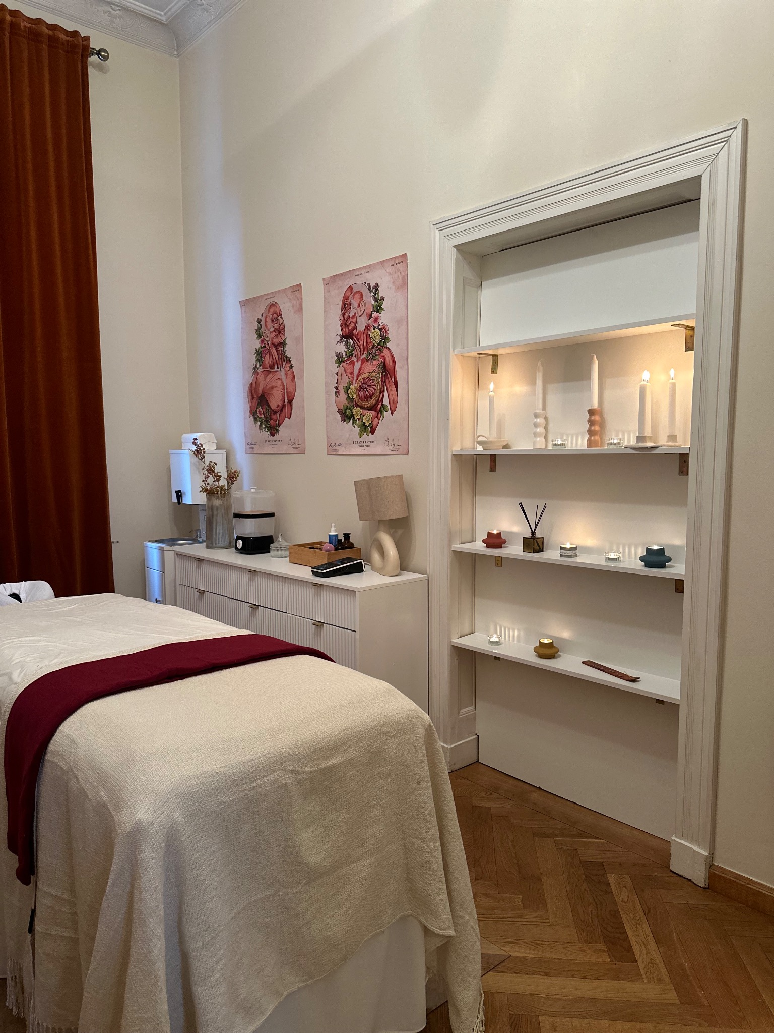 Przytulne wnętrze gabinetu z łóżkiem do zabiegów, białymi meblami, dekoracyjnymi świecami w półce i plakatami anatomicznymi na ścianie.