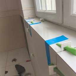 KVB CONSTRUCTION KAMIL VIP BUD Kamil Damps - Białe płytki układane przy oknie w łazience, z użyciem niebieskiej taśmy i zielonych klinów poziomujących płytki.