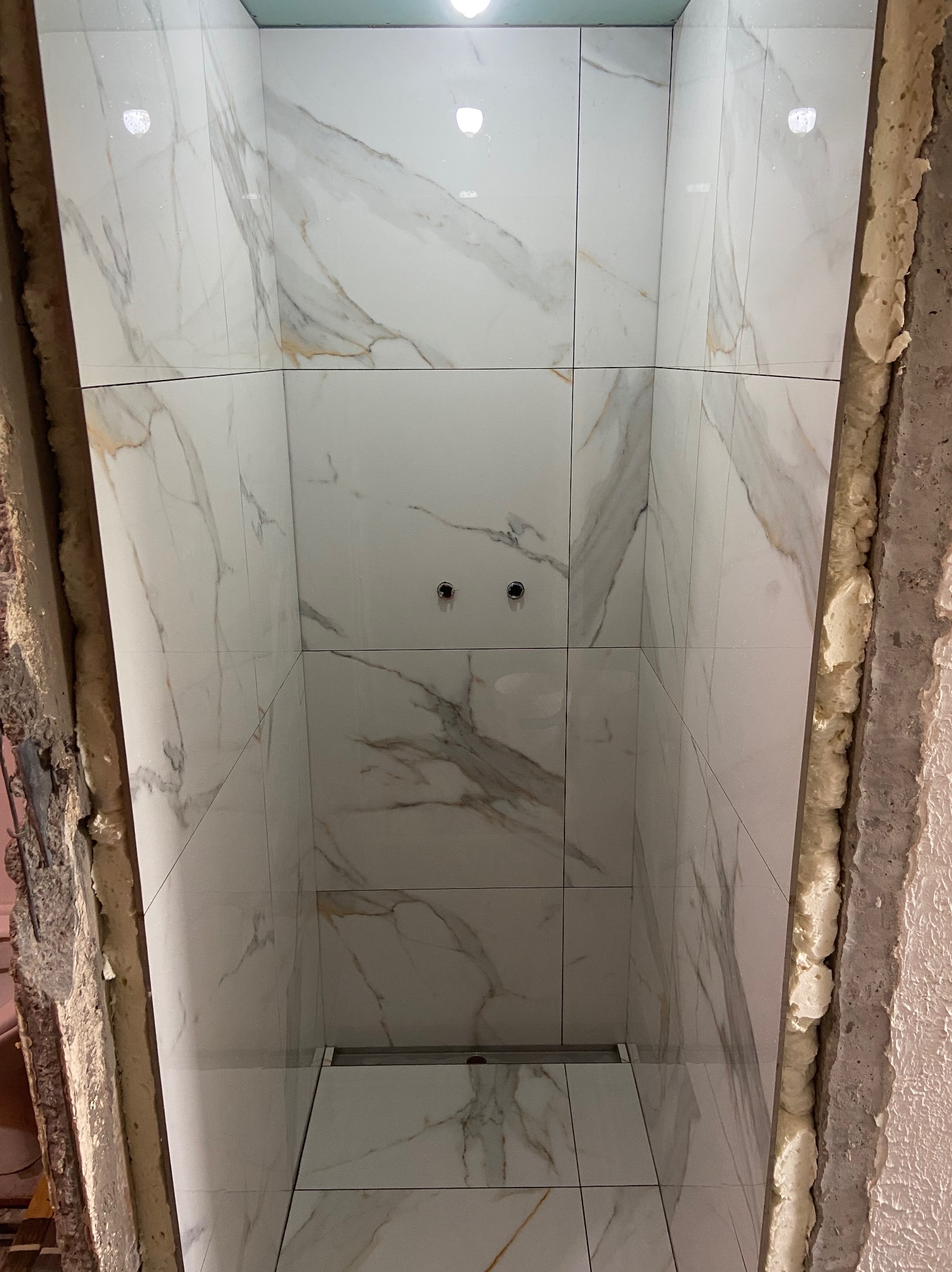 Wnętrze kabiny prysznicowej w trakcie wykańczania, widoczne białe płytki imitujące marmur na ścianach, zamontowany odpływ liniowy w podłodze oraz pianka montażowa wokół krawędzi.