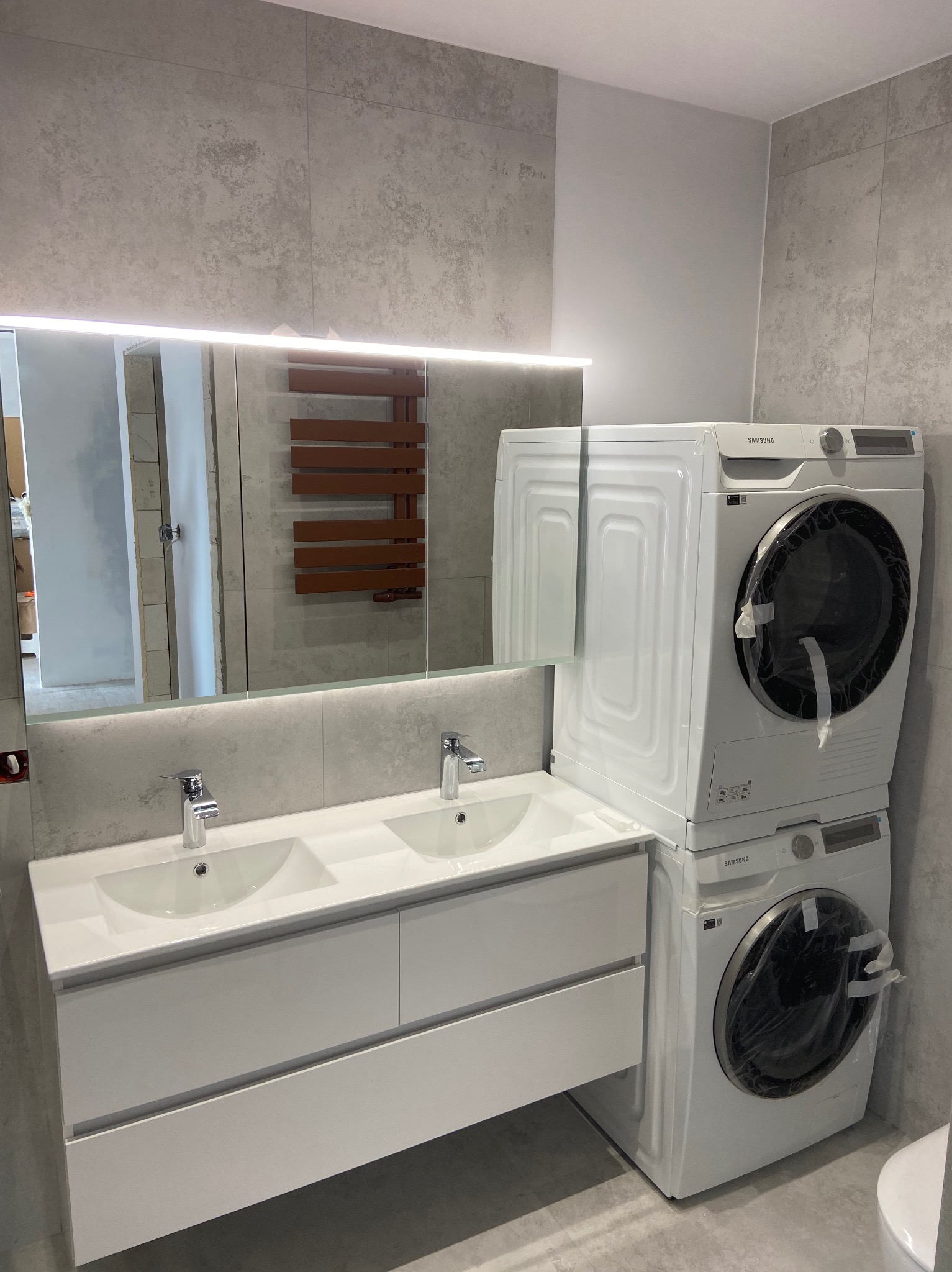 Nowoczesna łazienka w szarościach z podwójną umywalką na szafce i pralko-suszarką Samsung ustawioną w kolumnie, widoczne zabezpieczenia transportowe na sprzęcie.