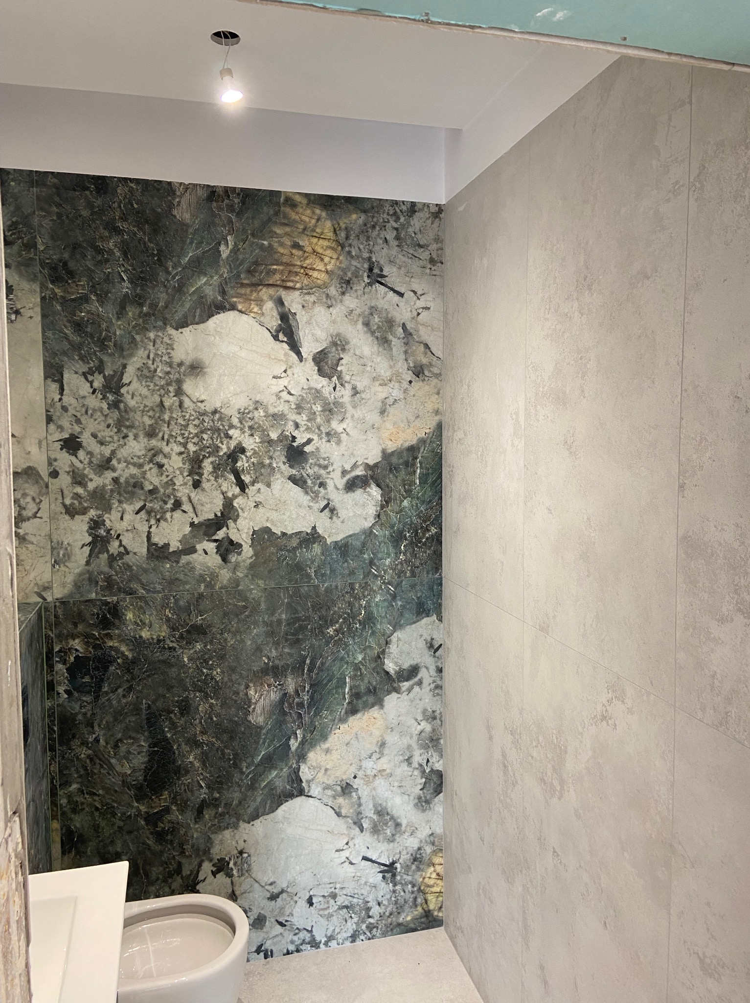 Płytki imitujące marmur w odcieniach zieleni i szarości na ścianie łazienki, zestawione z gładkimi, beżowymi płytkami na sąsiedniej ścianie, widoczna biała miska toaletowa i sufit z punktowym...