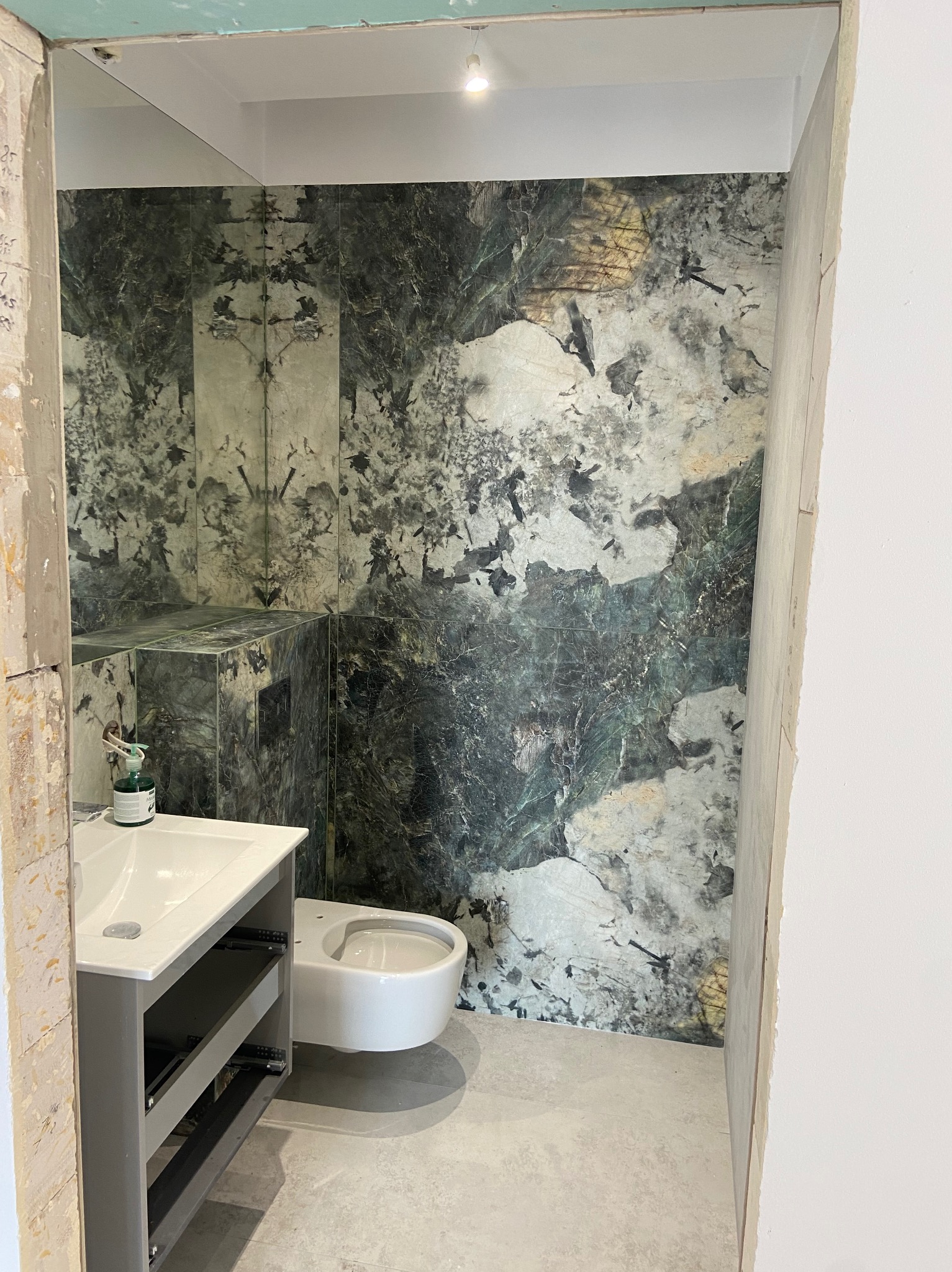 Łazienka w trakcie remontu z płytkami imitującymi marmur na ścianach, wisząca toaleta i szafka pod umywalkę bez frontów szuflad.