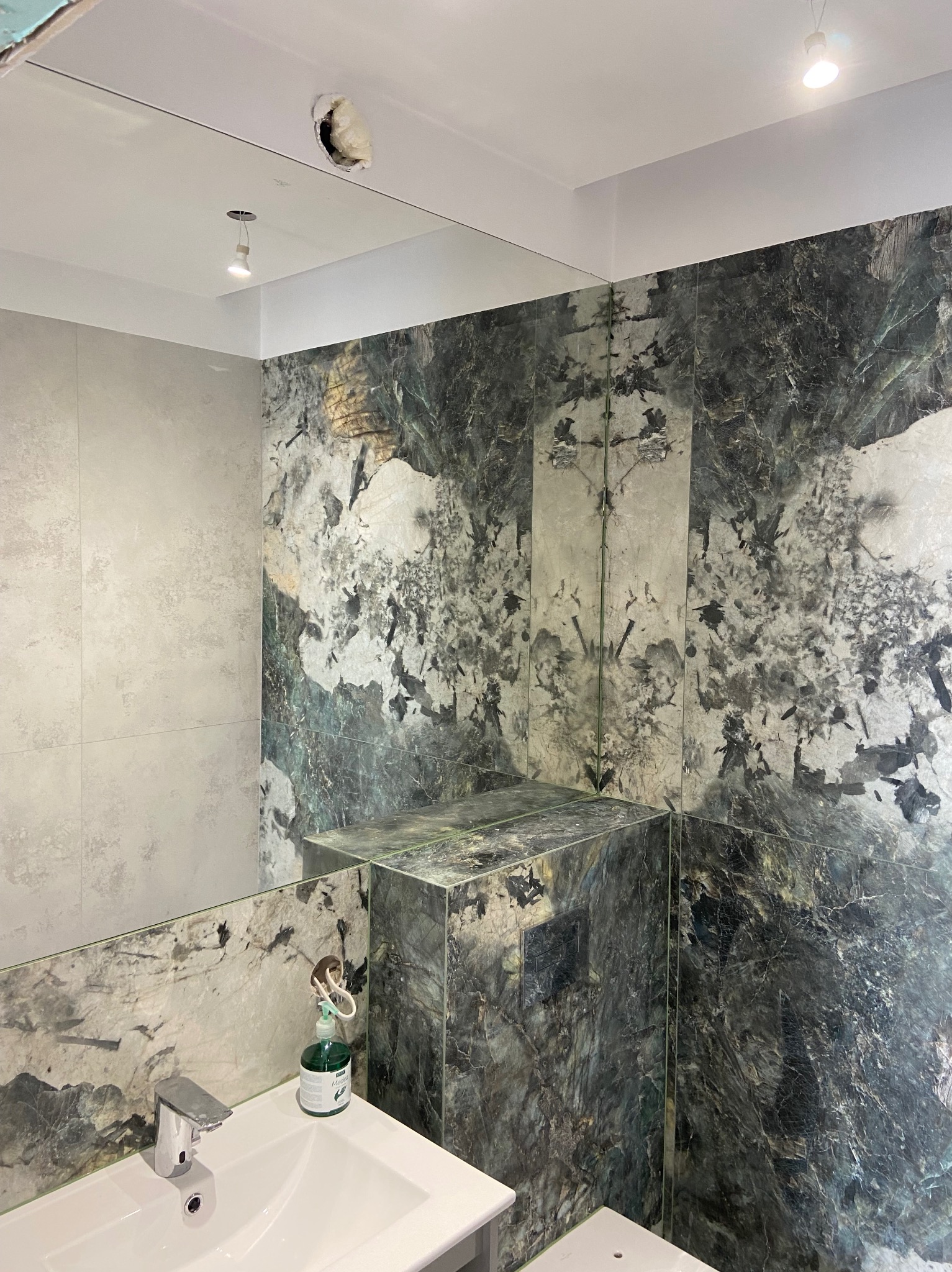 Nowoczesna łazienka z płytkami imitującymi marmur w odcieniach szarości i zieleni, widoczne umywalka z baterią, lustro oraz otwór w suficie z wystającą izolacją.