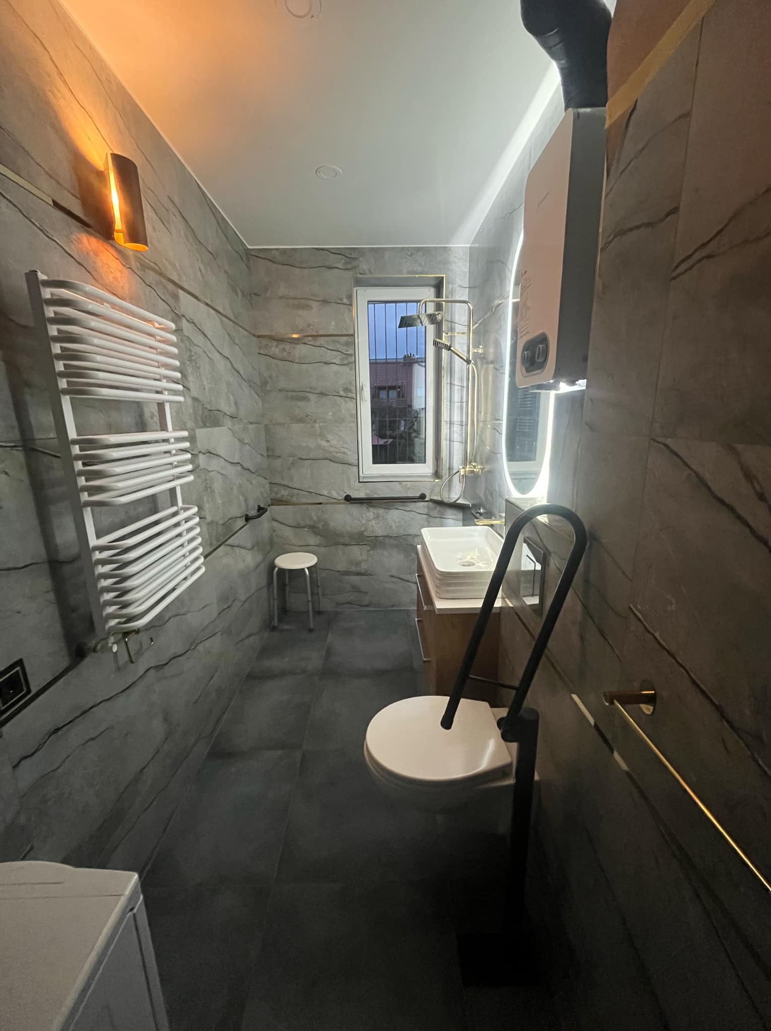 Nowoczesna łazienka z szarymi płytkami imitującymi kamień, podświetlanym lustrem, białym grzejnikiem drabinkowym, toaletą z uchwytem dla niepełnosprawnych i oświetleniem punktowym.