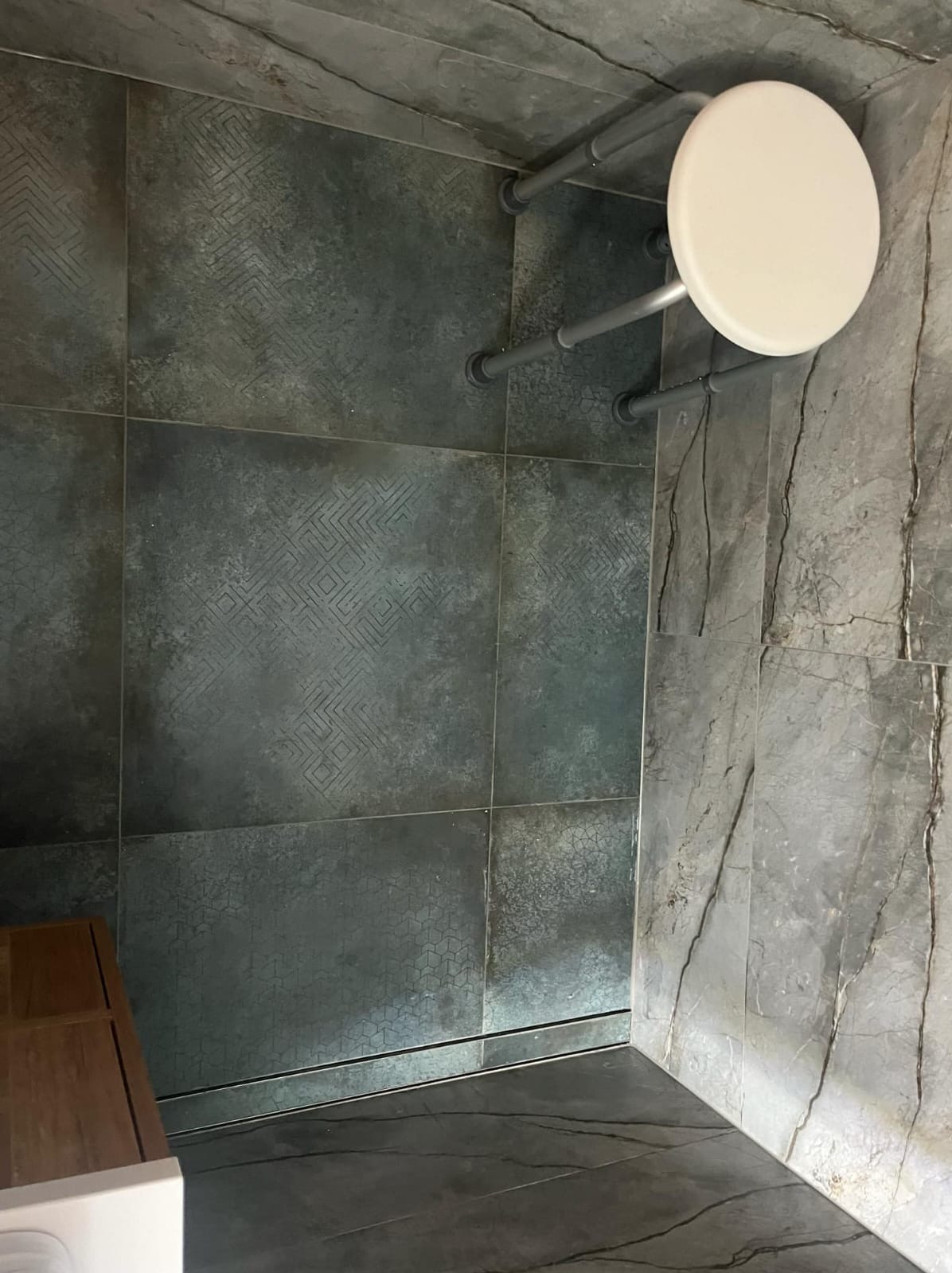 Wnętrze kabiny prysznicowej wyłożonej ciemnymi, wzorzystymi płytkami ceramicznymi, z zamontowanym siedziskiem na metalowym stelażu i odpływem liniowym w podłodze.