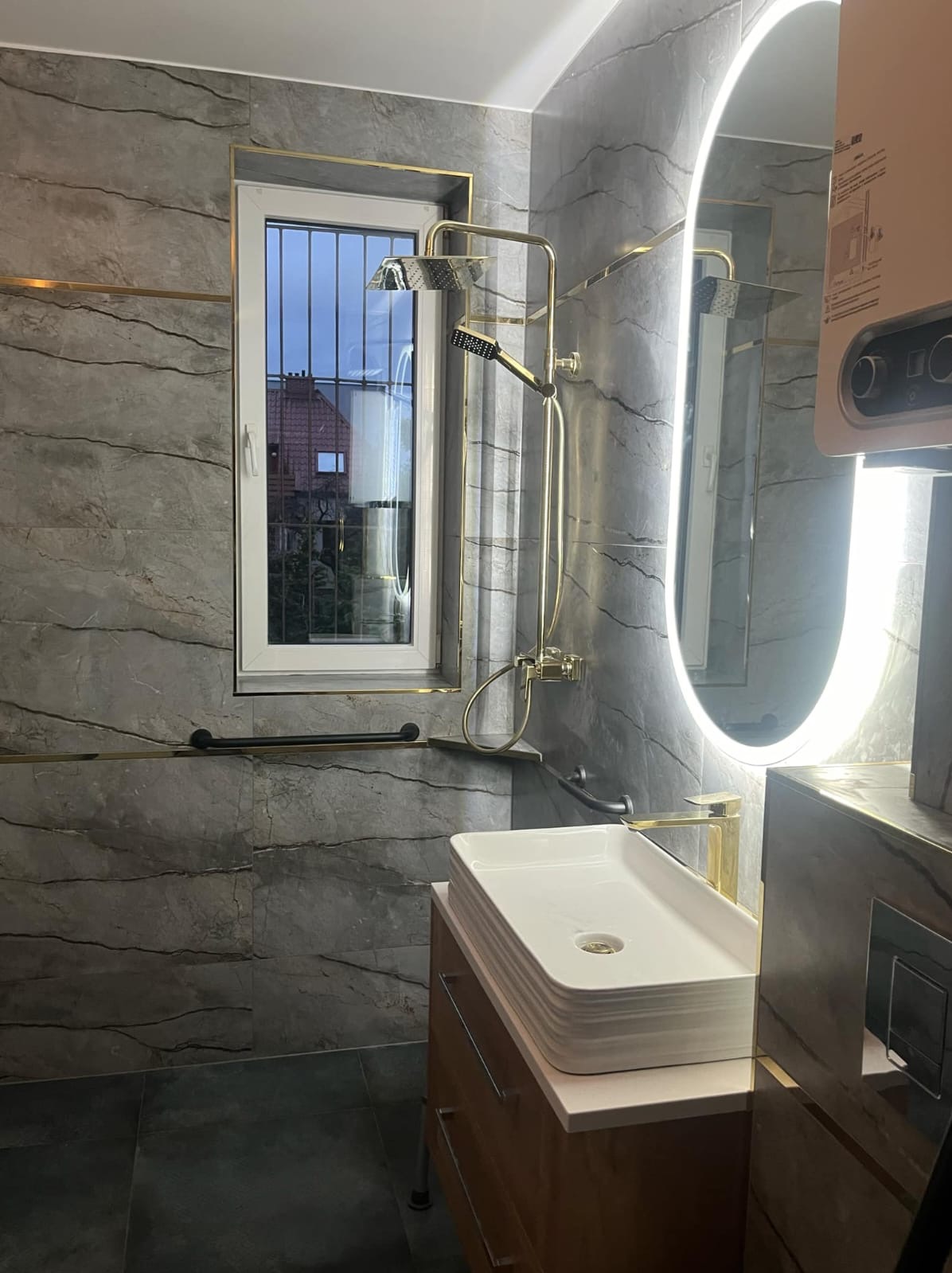 Wykończona łazienka z szarymi płytkami imitującymi marmur, złotymi akcentami, owalnym lustrem z podświetleniem LED oraz białą, prostokątną umywalką na drewnianej szafce.