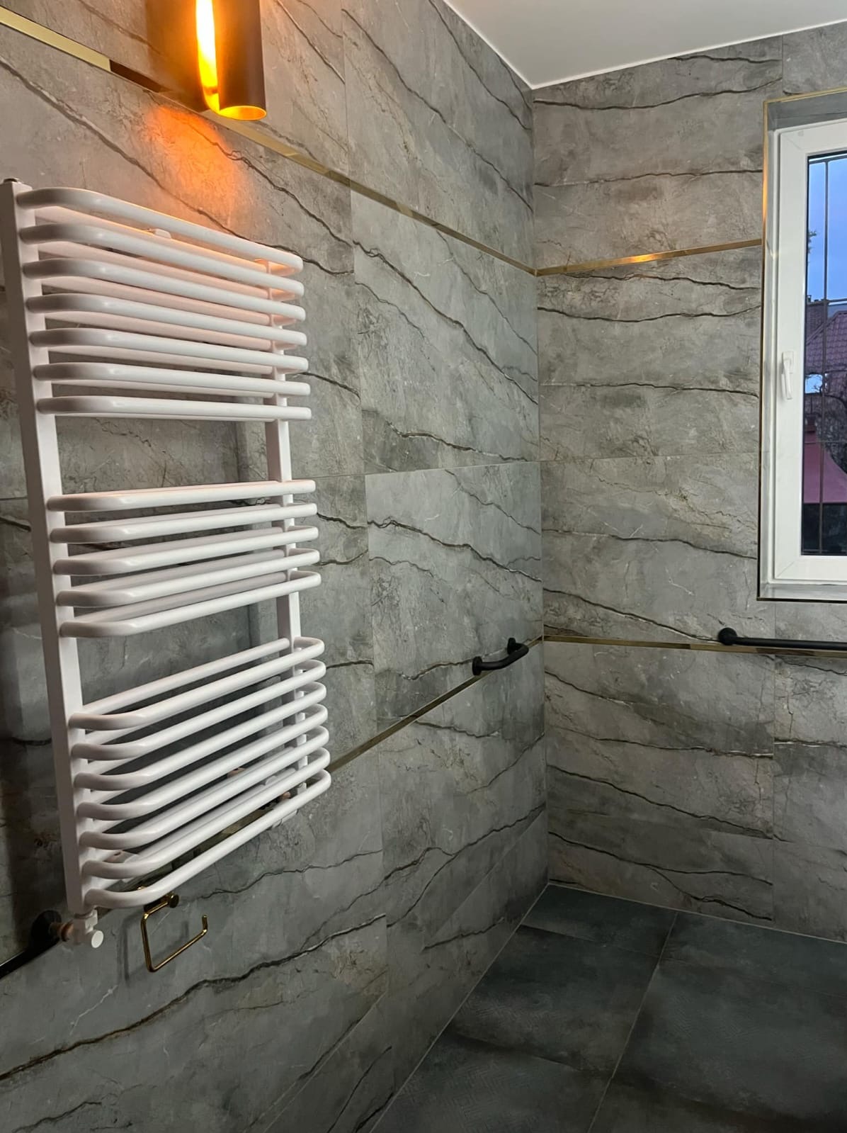 Wykończona łazienka z grzejnikiem drabinkowym, płytki imitujące marmur z delikatnymi złotymi listwami, czarny uchwyt na ręczniki i oświetlenie punktowe.