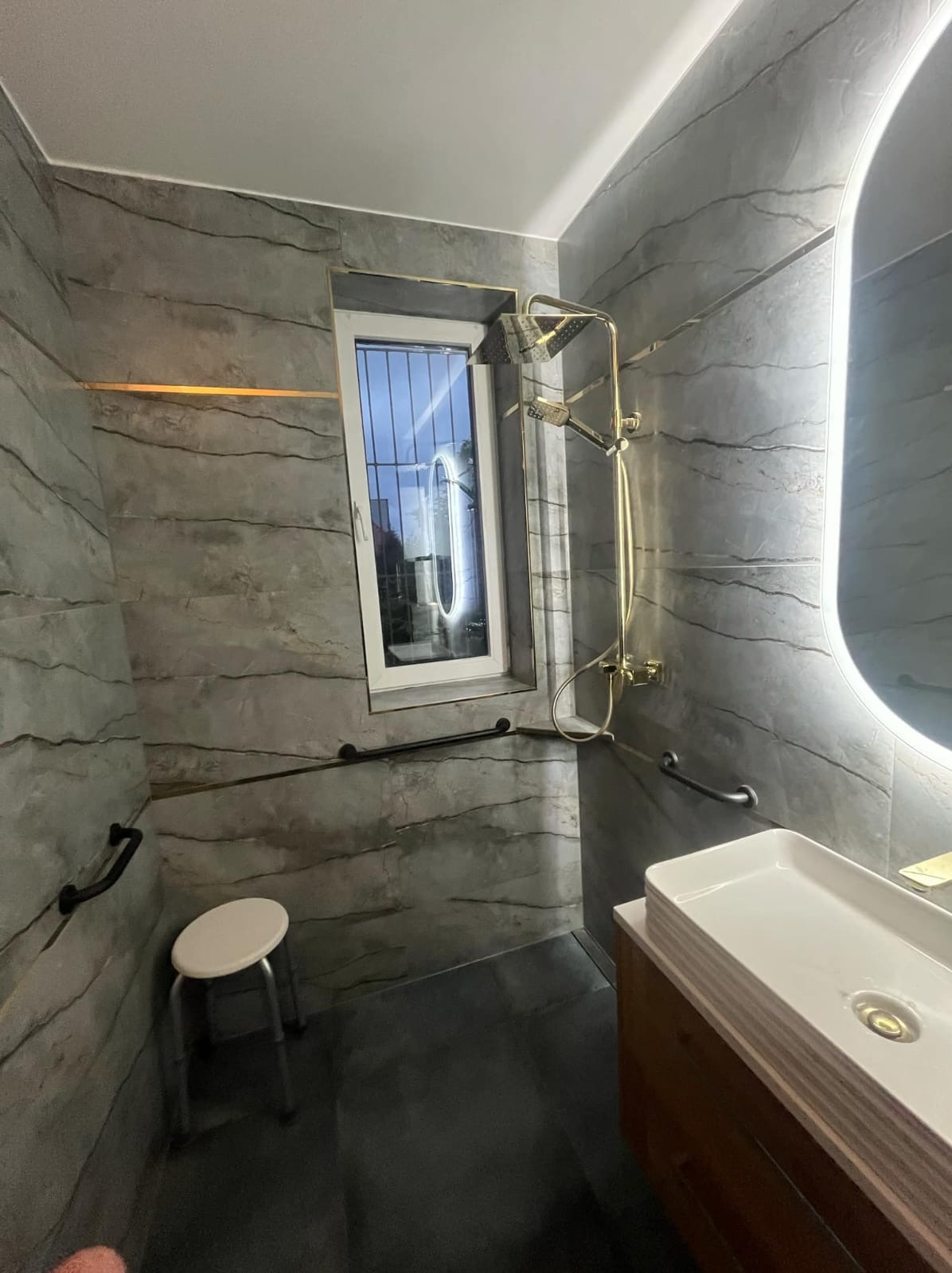 Szara łazienka z marmurowymi płytkami, złotą armaturą i podświetlanym lustrem, dodatkowe uchwyty dla niepełnosprawnych, małe okno z widokiem na zewnątrz, taboret.