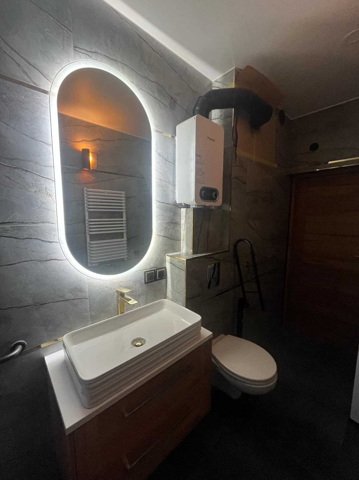 Nowoczesna łazienka z szarą, kamienną okładziną, owalnym lustrem z podświetleniem LED, prostokątną umywalką nablatową z złotą baterią, białym bojlerem i toaletą z czarną poręczą.