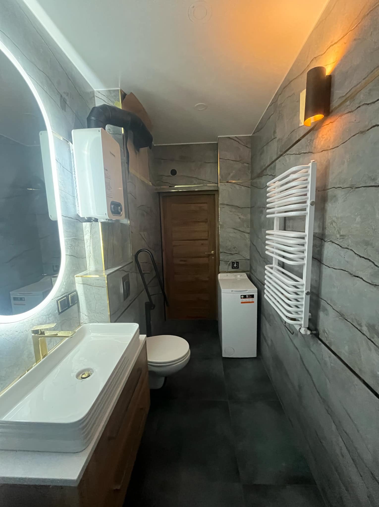 Nowoczesna łazienka z umywalką nablatową z złotą armaturą, podświetlanym lustrem, grzejnikiem drabinkowym, pralką i piecem gazowym na ścianie z szarych płytek imitujących kamień.