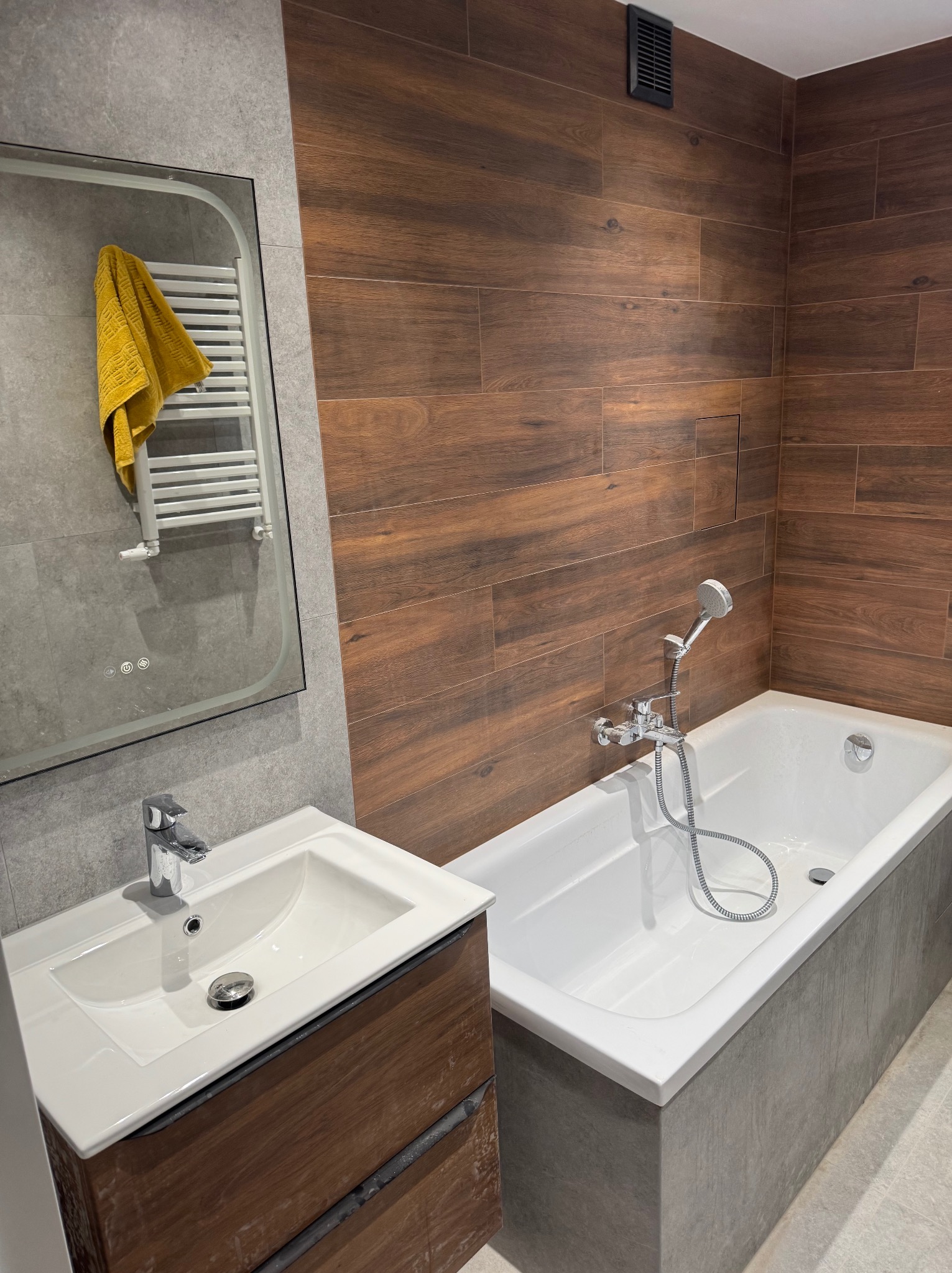 Nowoczesna łazienka z drewnopodobnymi płytkami na ścianie, białą wanną i umywalką z szafką z ciemnego drewna, lustro z podświetleniem i grzejnikiem drabinkowym z powieszonym żółtym ręcznikiem.