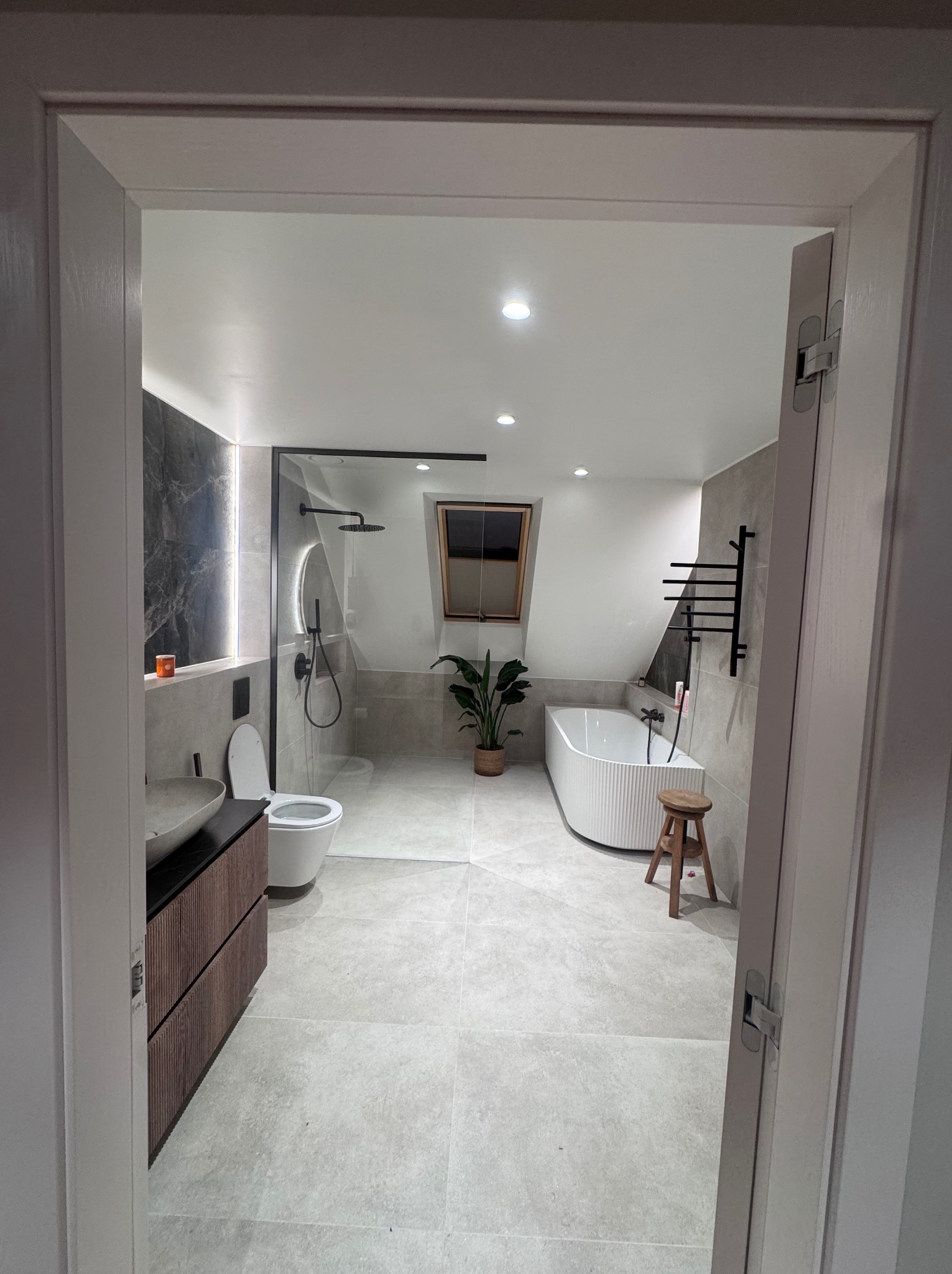 Nowoczesna łazienka na poddaszu z kabiną prysznicową typu walk-in, wolnostojącą wanną i drewnianą szafką pod umywalką. Widok zza otwartych drzwi.