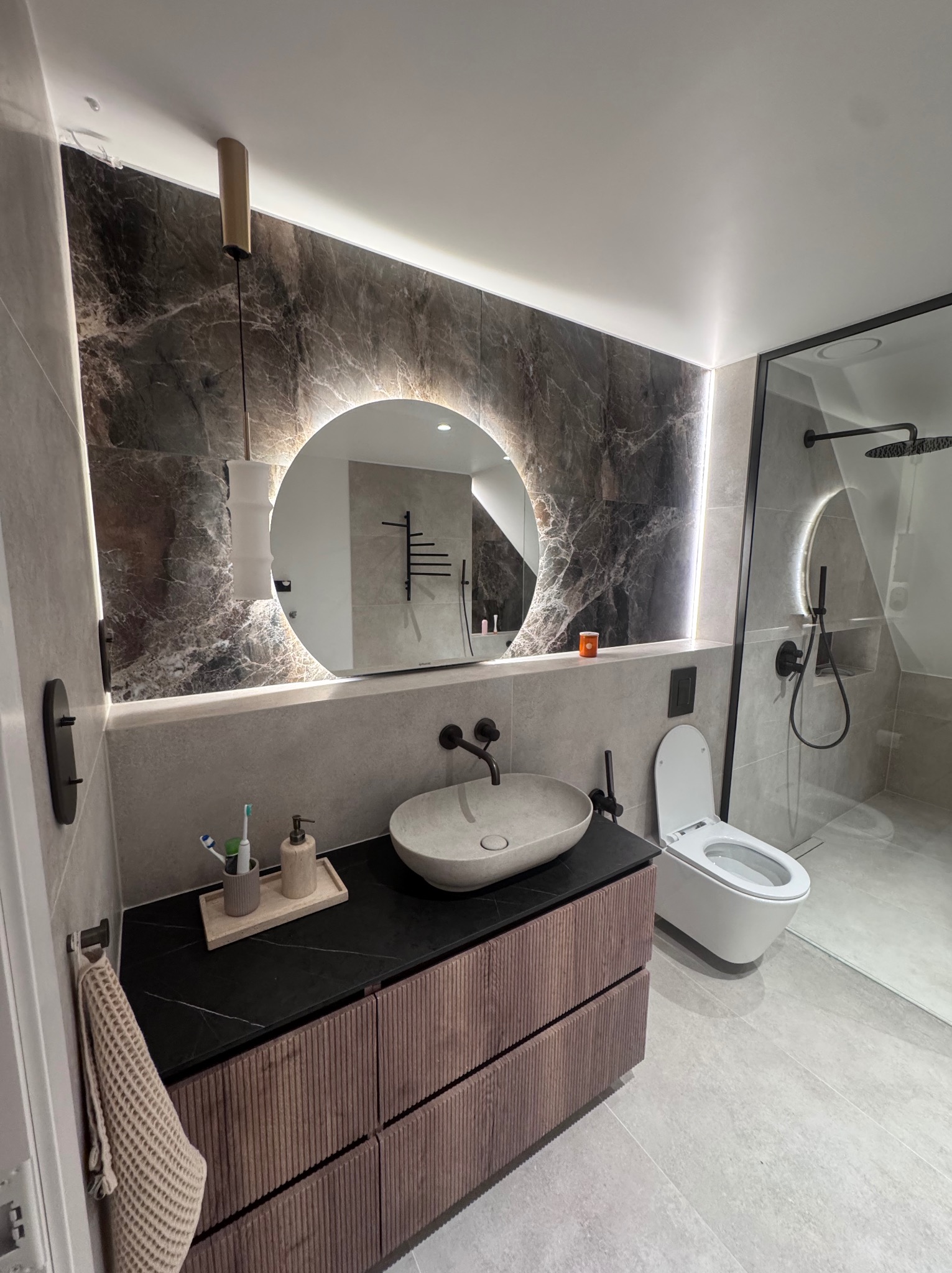 Nowoczesna łazienka z okrągłym lustrem LED i marmurową ścianą w Gdyni. Umywalka nablatowa na szafce z frezowanymi frontami. Kabina prysznicowa typu walk-in.