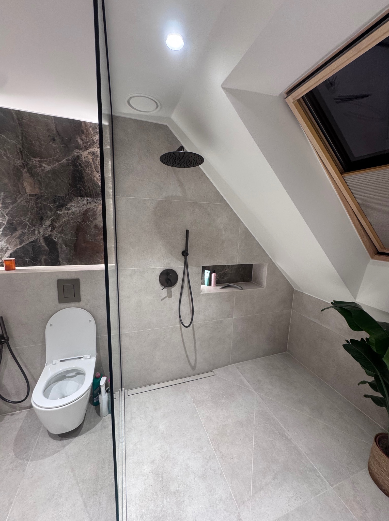 Nowoczesna łazienka na poddaszu z szarą glazurą, kabiną typu walk-in, toaletą i oknem dachowym. W tle marmurowa ściana i roślina doniczkowa.