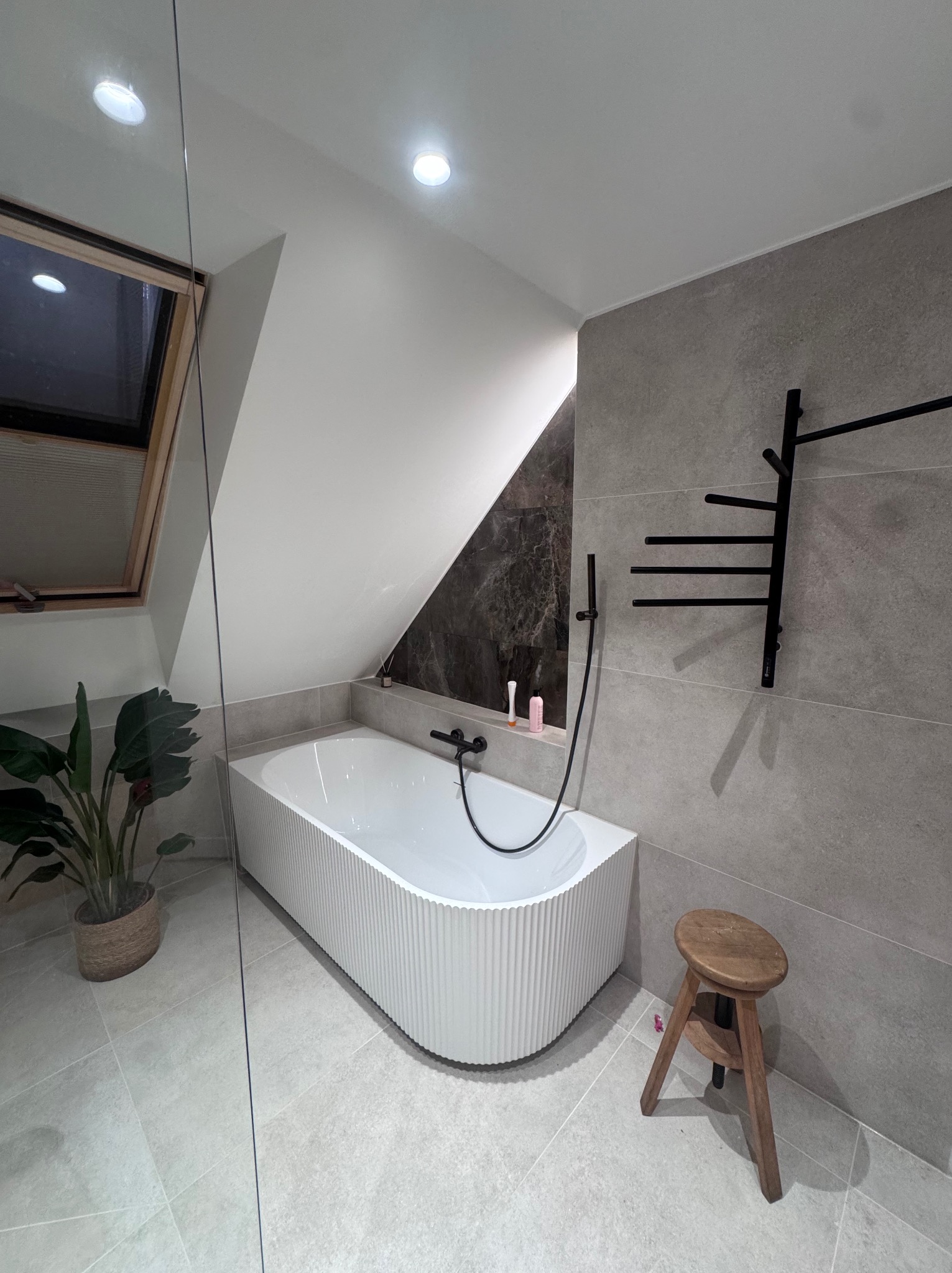 Nowoczesna łazienka na poddaszu z białą wanną o żłobkowanej fakturze, szarą płytką i drewnianym stołkiem. Uwagę zwraca okno dachowe i czarny grzejnik drabinkowy.