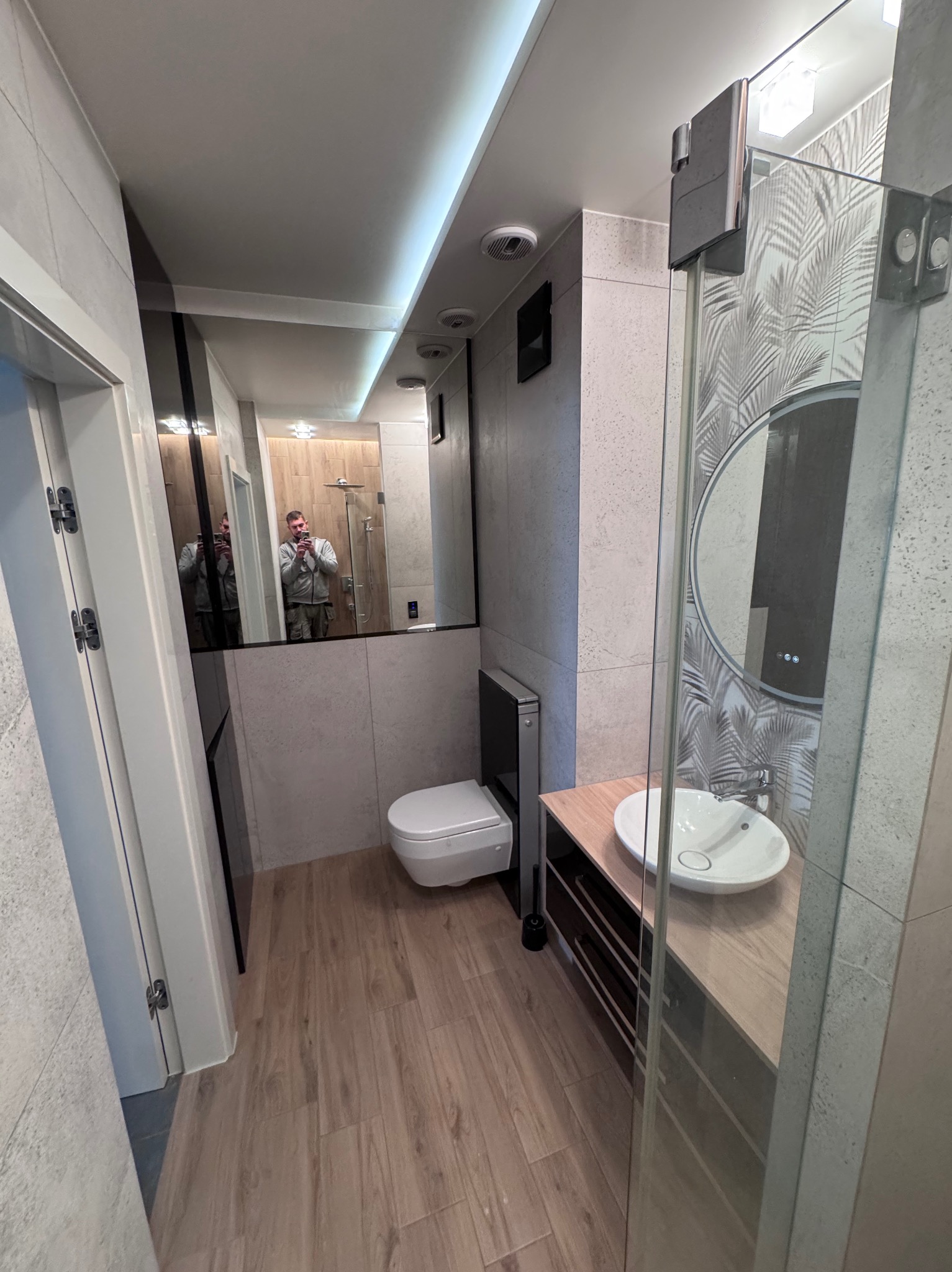 Nowoczesna łazienka w Gdyni po remoncie: drewnopodobne panele, szara glazura, kabina prysznicowa ze wzorem liści, okrągłe lustro i podwieszana toaleta.