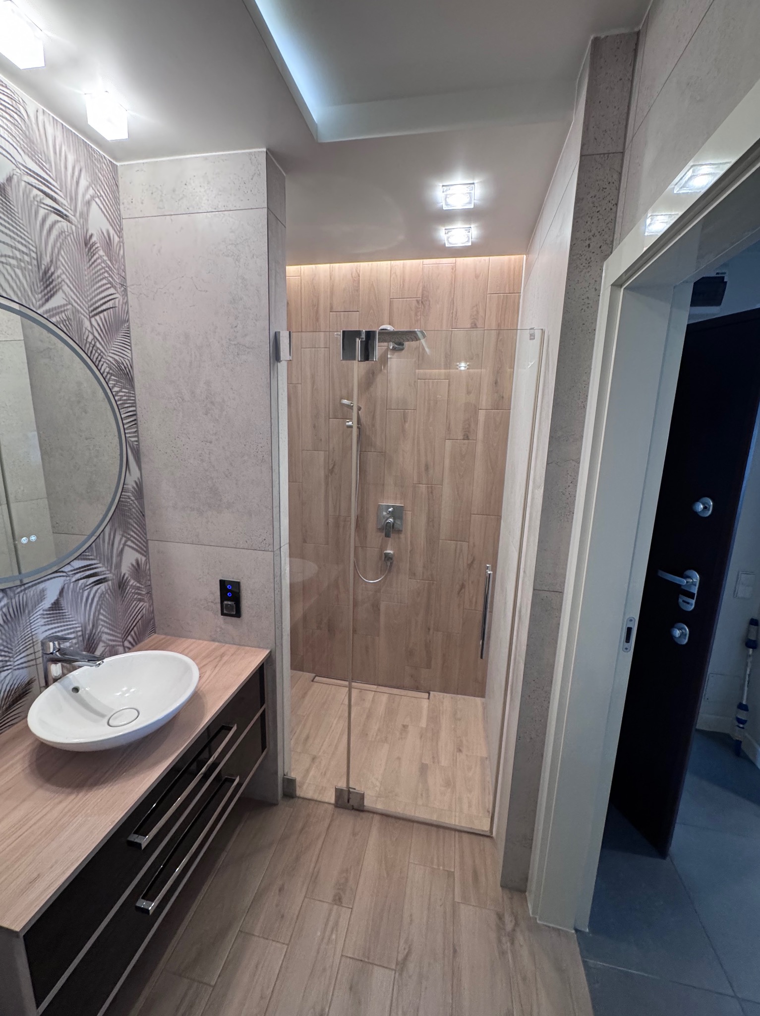 Nowoczesna łazienka po remoncie: kabina prysznicowa ze szklanymi drzwiami, umywalka nablatowa z szafką, lustro okrągłe i płytki imitujące drewno na ścianach i podłodze.