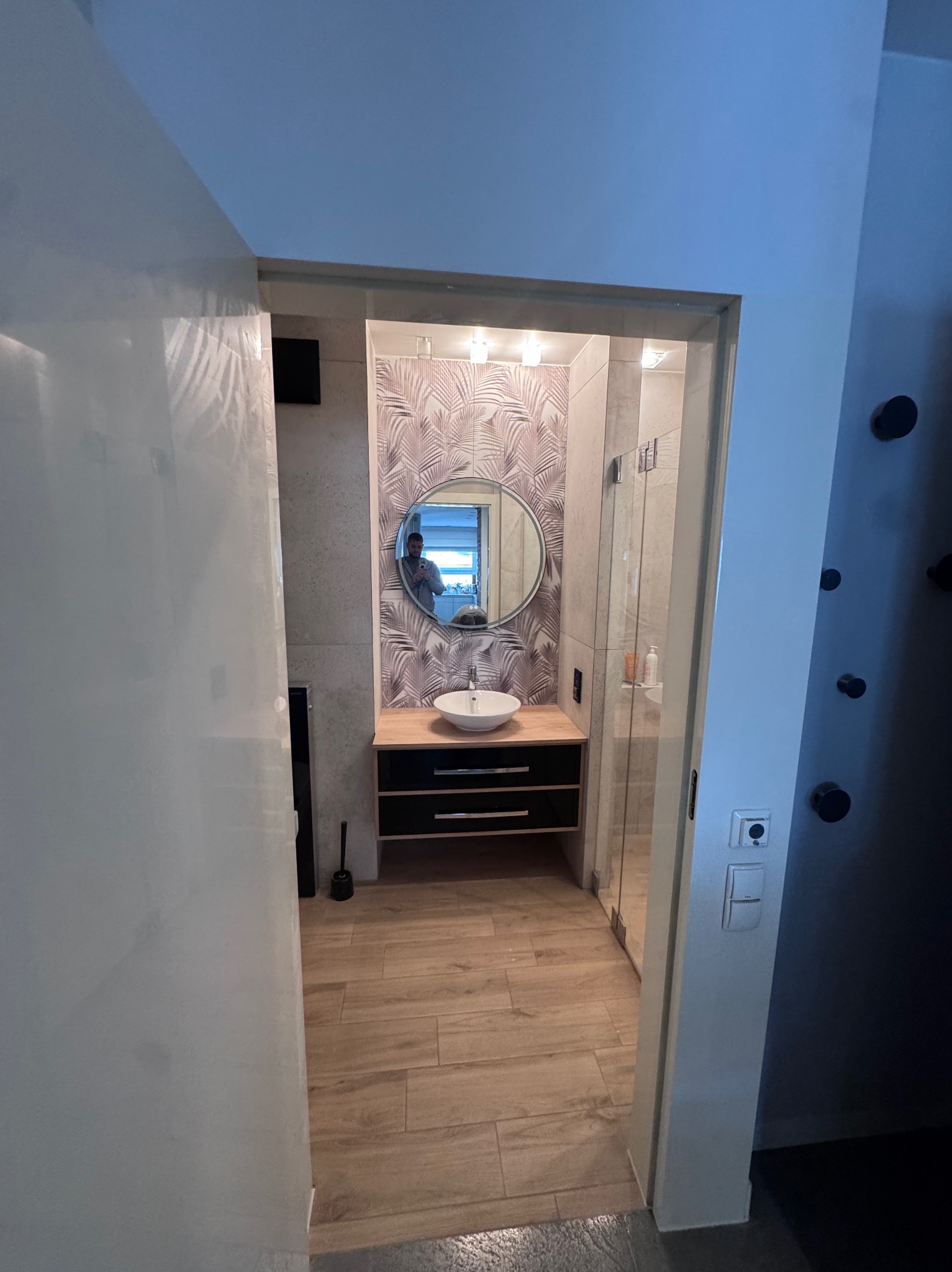 Nowoczesna łazienka z okrągłym lustrem i wzorzystą tapetą. Kabina prysznicowa ze szklanymi drzwiami. Drewnopodobne płytki na podłodze. Widok z korytarza.