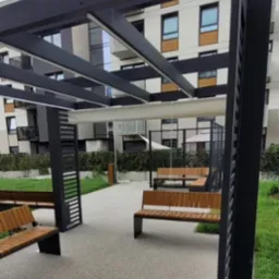 Nowoczesna aranżacja przestrzeni publicznej w Świdnicy: szara nawierzchnia z żywicy, drewniane ławki z czarnymi metalowymi nogami pod ażurową, czarną pergolą, w tle budynek mieszkalny.