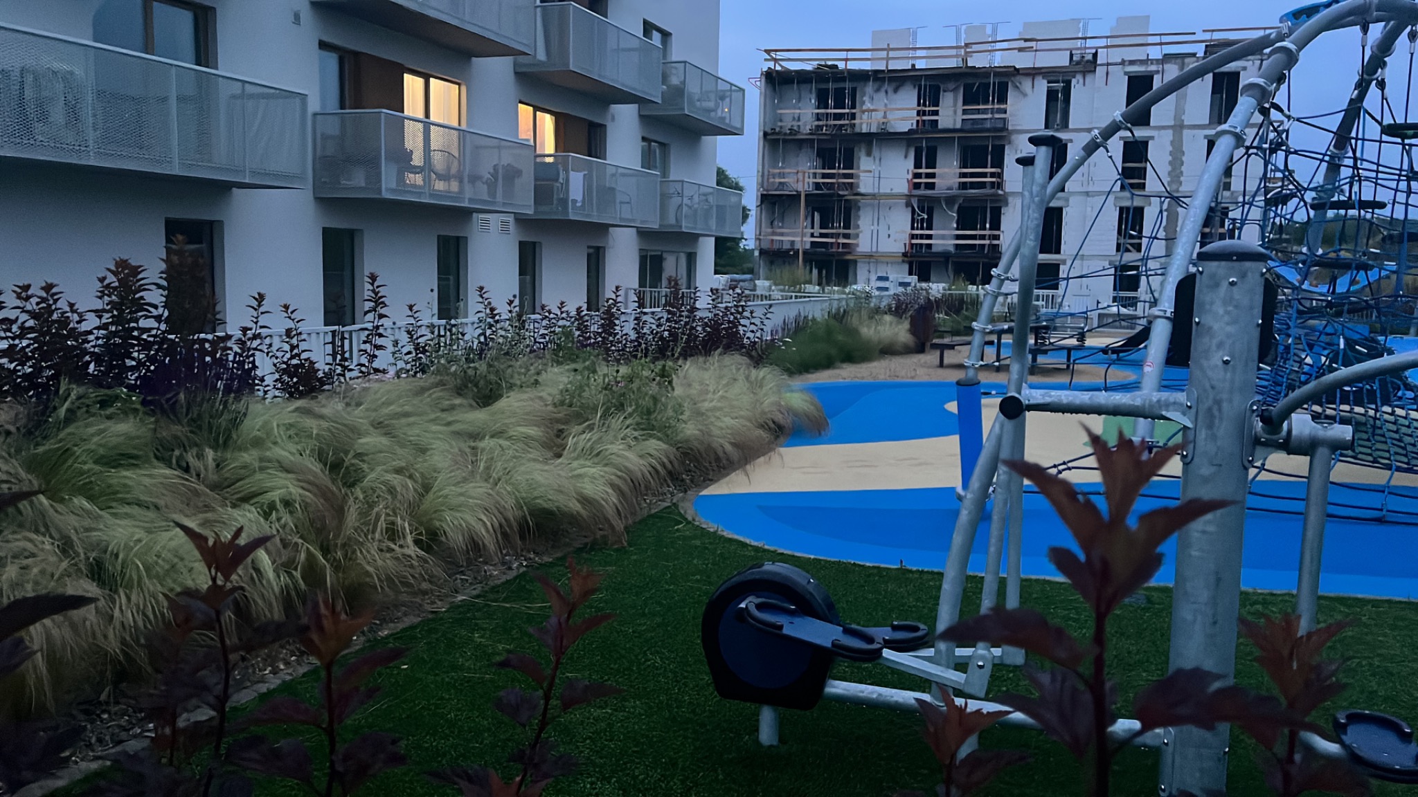 Nowoczesny plac zabaw z elementami do ćwiczeń na tle budynku mieszkalnego i konstrukcji w trakcie budowy, z dekoracyjnymi trawami i roślinami na pierwszym planie, sfotografowany o zmierzchu.