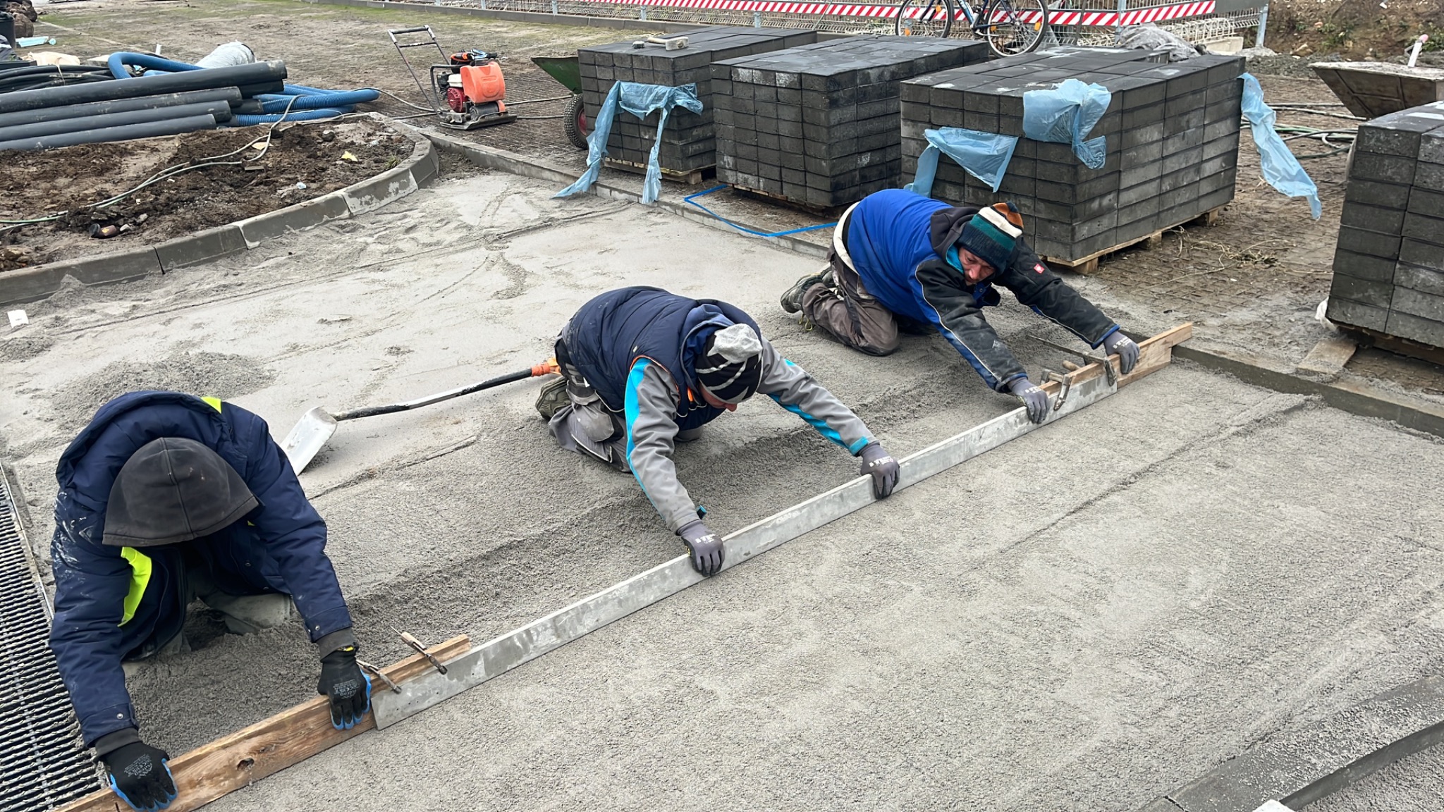 Trzech pracowników na kolanach wyrównuje piasek podłoża pod kostkę brukową za pomocą długiej poziomicy i drewnianych łat, w tle palety z kostką, zagęszczarka i rury.