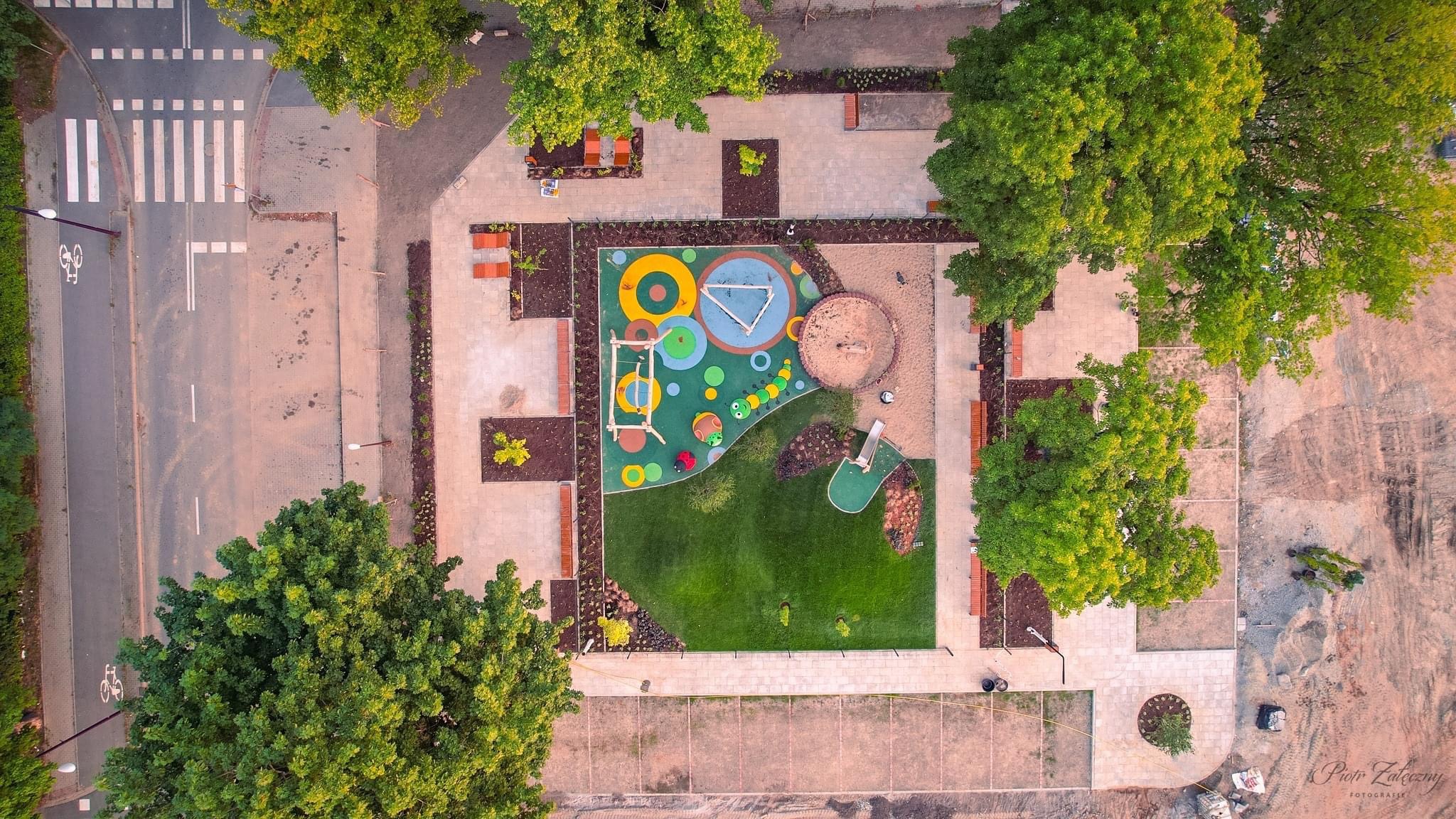 Widok z lotu ptaka na nowo wybudowany plac zabaw z kolorową nawierzchnią, piaskownicą, zjeżdżalnią, otoczony chodnikiem z kostki, zielenią i drzewami, sąsiadujący z ulicą i parkingiem.