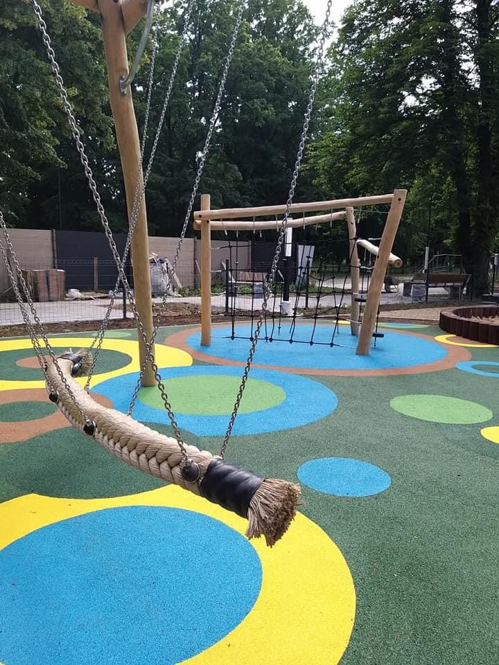 Huśtawka linowa na placu zabaw z kolorową nawierzchnią w kształcie kół, w tle konstrukcja do wspinaczki i drzewa.