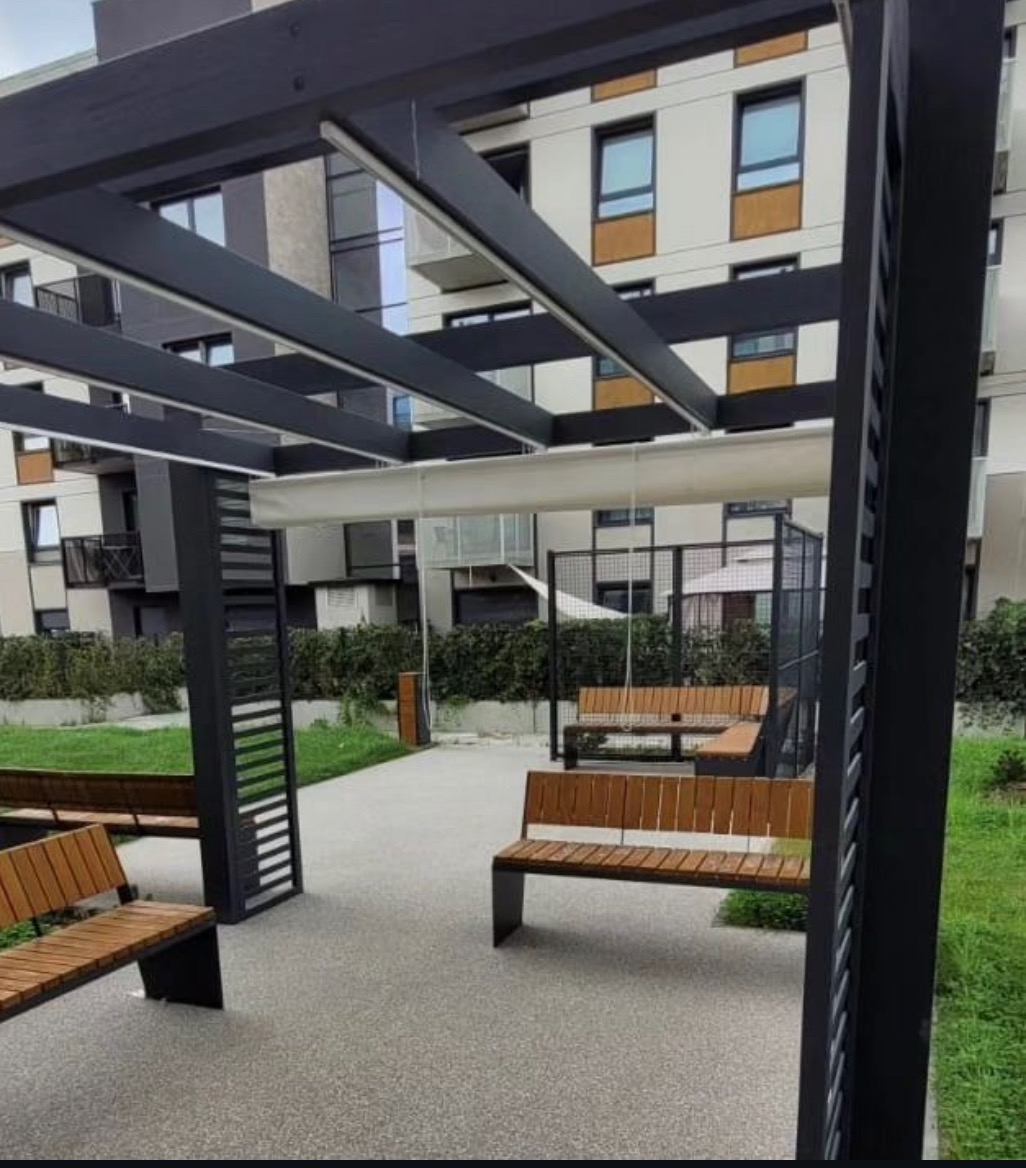 Nowoczesna aranżacja przestrzeni publicznej w Świdnicy: szara nawierzchnia z żywicy, drewniane ławki z czarnymi metalowymi nogami pod ażurową, czarną pergolą, w tle budynek mieszkalny.