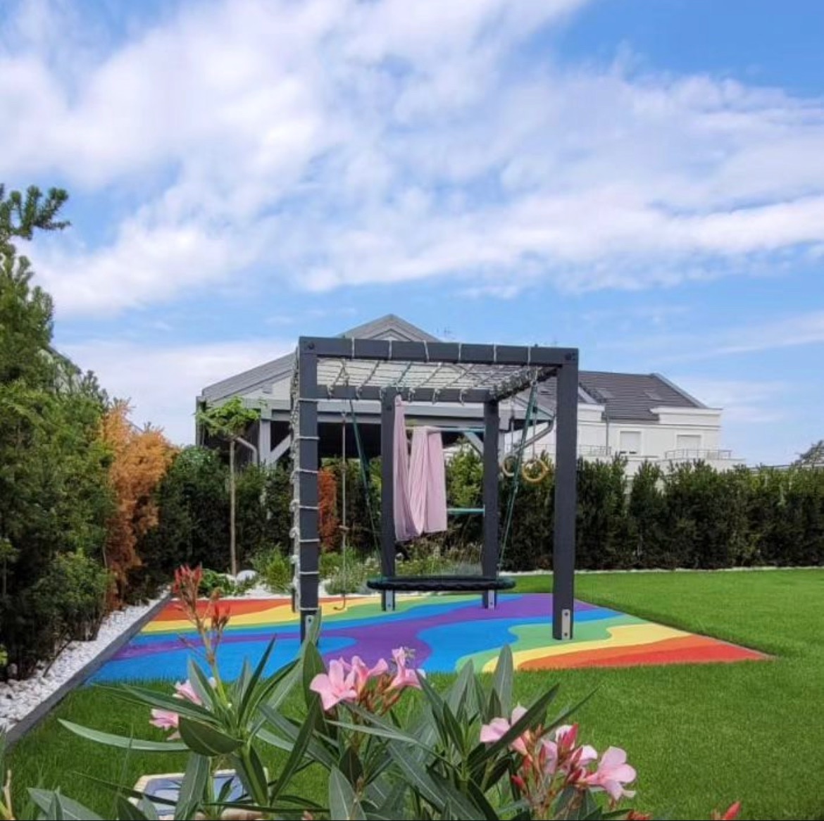 Kolorowa nawierzchnia placu zabaw z huśtawką na tle zadbanego ogrodu i domu.