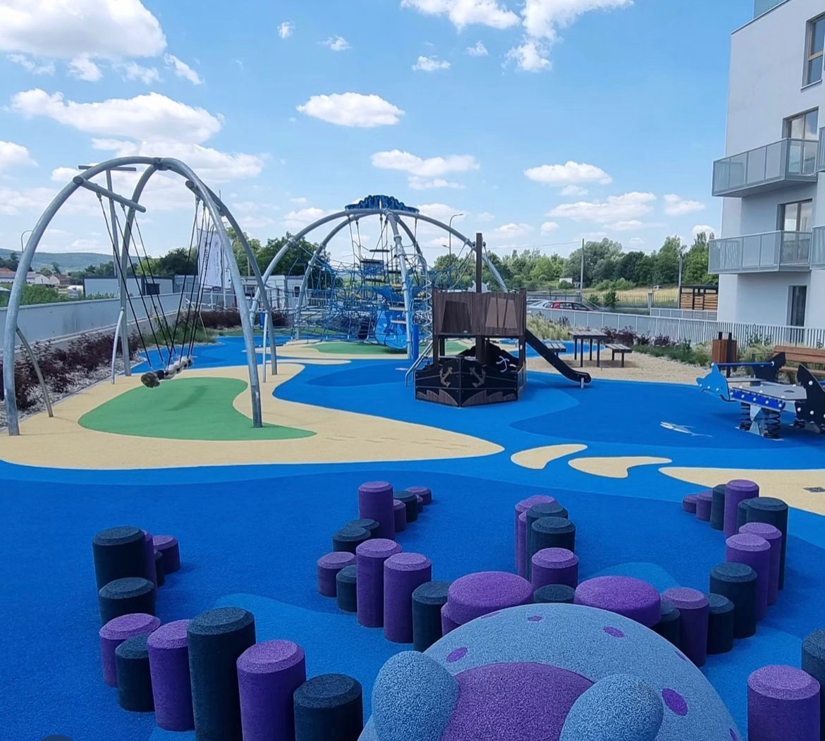 Kolorowy plac zabaw z huśtawkami, konstrukcjami linowymi i elementami w kształcie grzybków, otoczony budynkami mieszkalnymi w pogodny dzień.