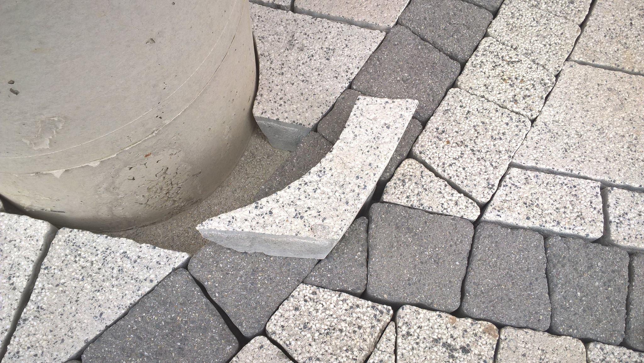 Niedokończona nawierzchnia z kostki granitowej w odcieniach szarości i bieli, z pojedynczym elementem w kształcie klina leżącym obok betonowego słupa.