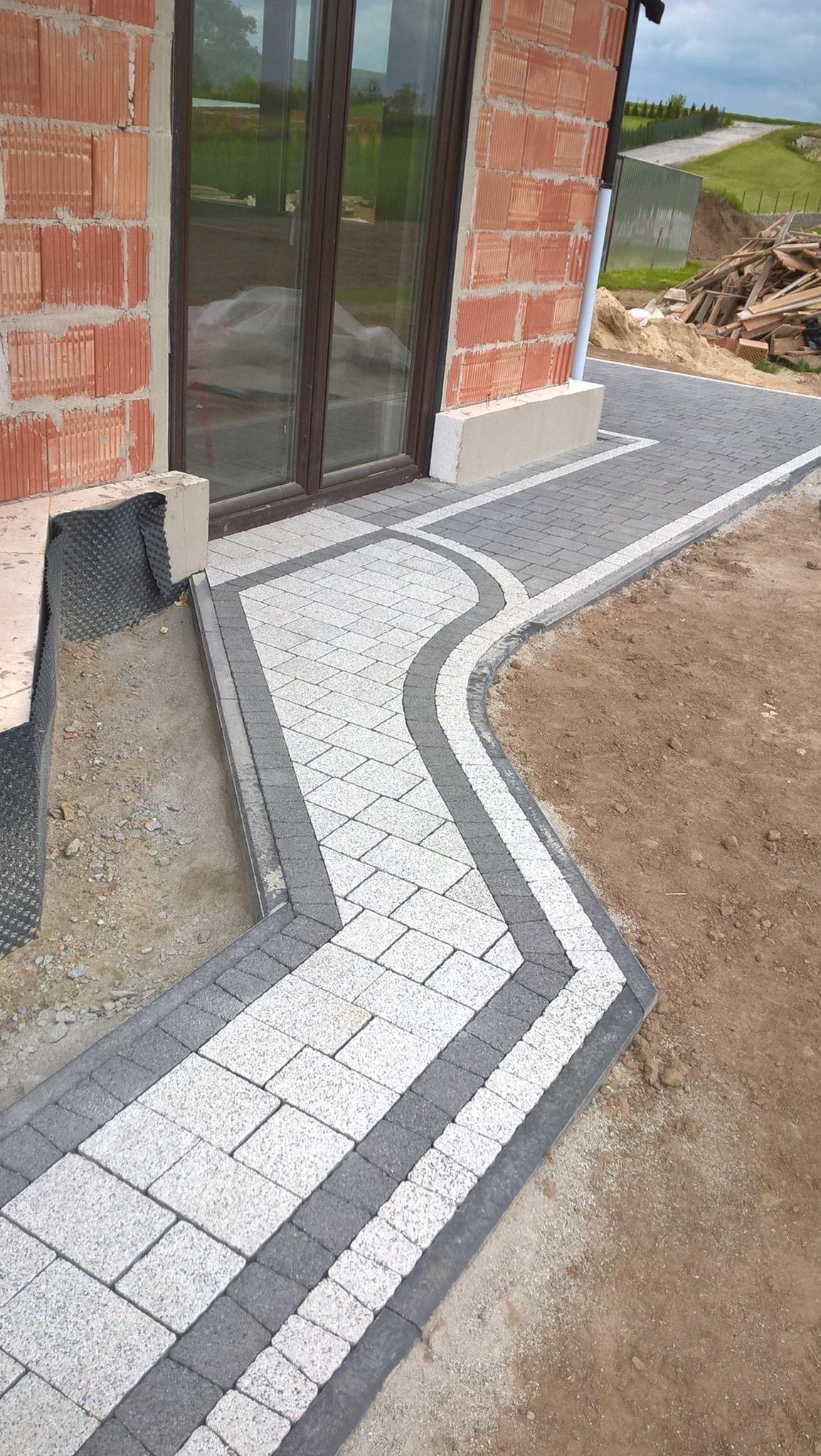 Świeżo ułożona ścieżka z jasnoszarej kostki brukowej z ciemnoszarym obramowaniem prowadząca do wejścia domu w stanie surowym, widoczne niedokończone fragmenty terenu.