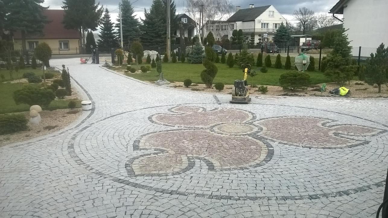 Ułożona z kostki granitowej nawierzchnia z dekoracyjnym motywem roślinnym w odcieniach szarości i beżu, widoczna ubijarka w tle oraz starannie zagospodarowany ogród z trawnikiem i ozdobnymi drzewami.