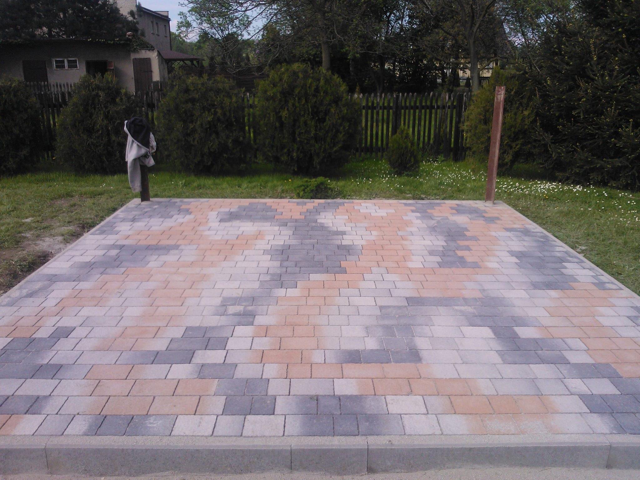 Ułożona nawierzchnia z kostki brukowej w odcieniach szarości i pomarańczu, obrzeża z szarego betonu, widok na ogród z tujami i drewnianym płotem w tle.