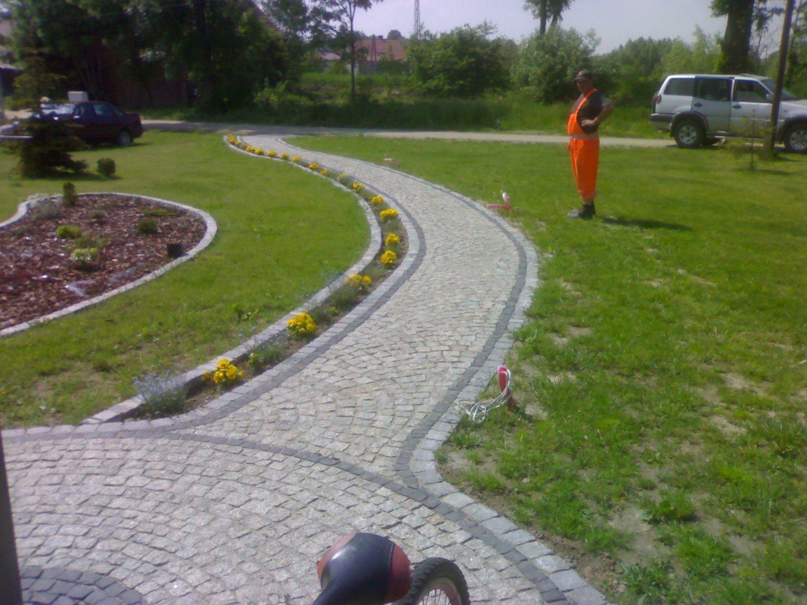 Kręta ścieżka z jasnej kostki granitowej z obramowaniem, obsadzona żółtymi kwiatami, prowadząca przez trawnik w kierunku domu, z pracownikiem w pomarańczowym kombinezonie stojącym obok i samochodem...
