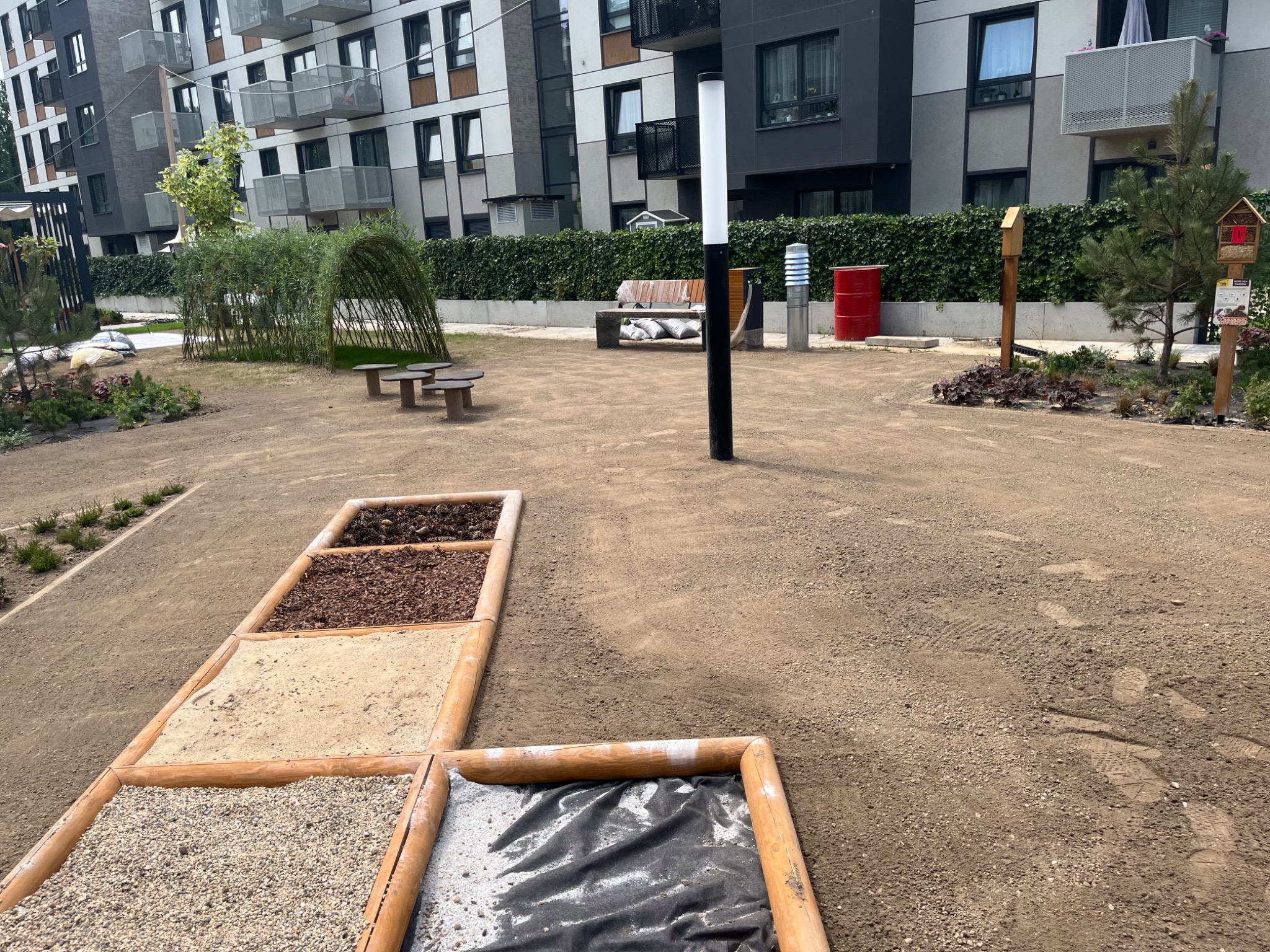 Nowoczesny plac zabaw z sensoryczną ścieżką z piasku, żwiru, kory i membrany, na tle zielonego żywopłotu i budynków mieszkalnych.