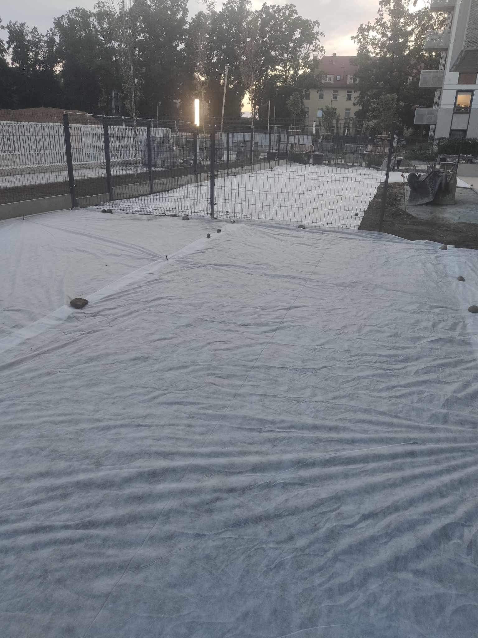 Przygotowanie terenu pod budowę: geowłóknina rozłożona na gruncie, zabezpieczona kamieniami, w tle ogrodzenie i budynek.