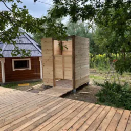 Drewniany taras prowadzący do szatni ogrodowej z surowego drewna, widok na mały domek i zieleń w tle.