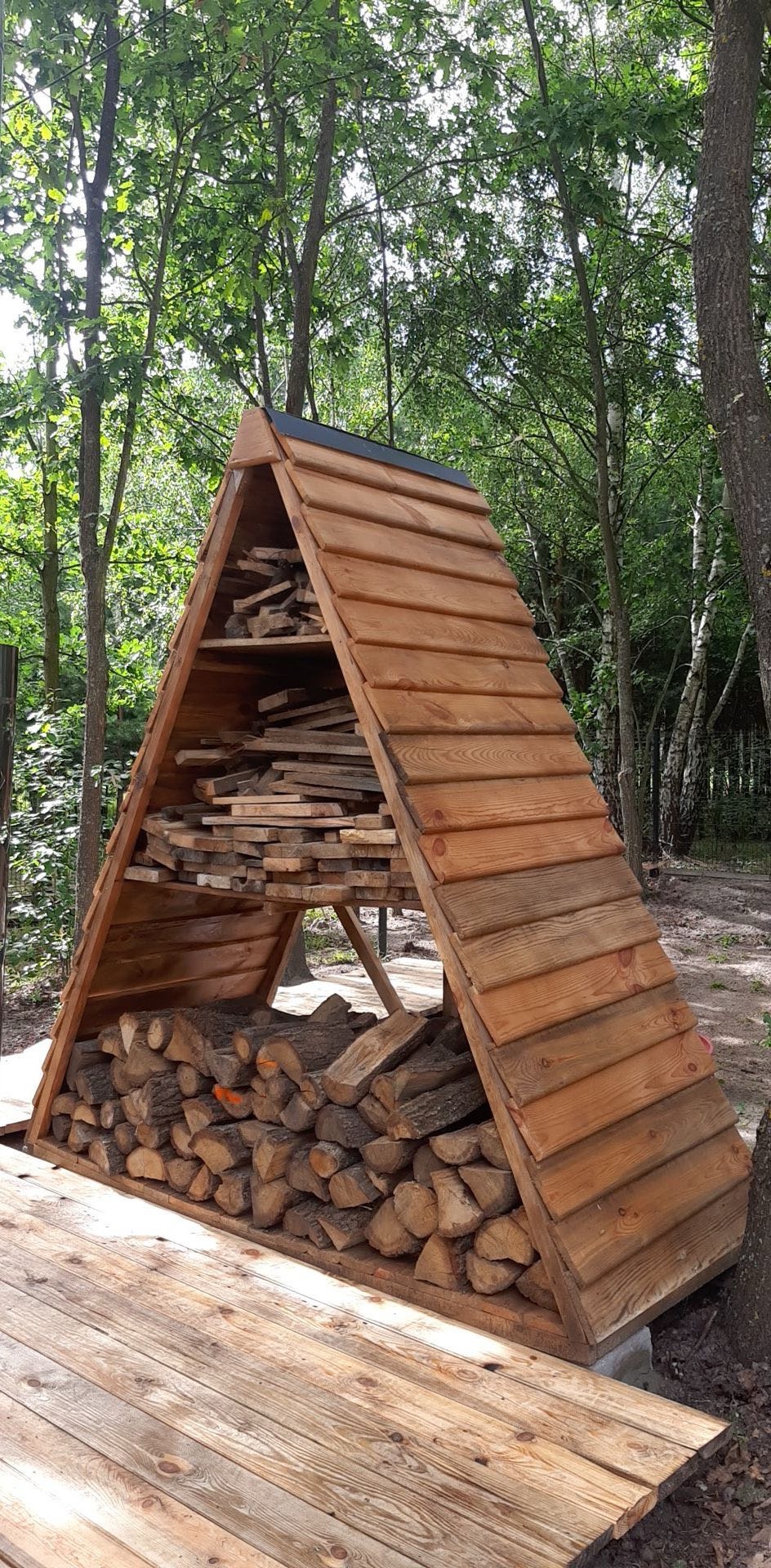 Drewniana, trójkątna konstrukcja z dachem, wypełniona pociętym drewnem i deskami, umieszczona na drewnianym podeście w otoczeniu drzew.