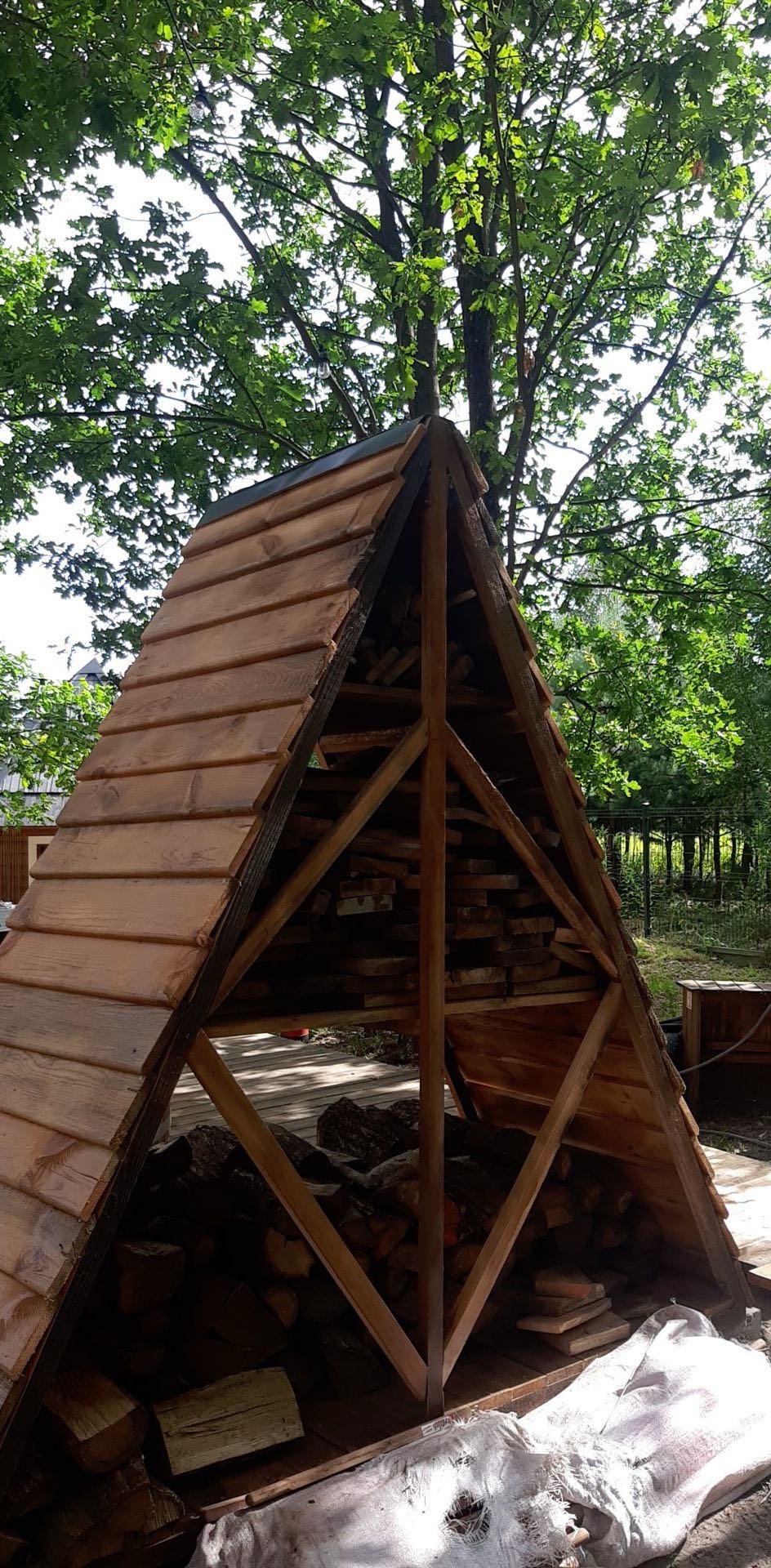 Drewniana konstrukcja w kształcie piramidy, zadaszona gontem, służąca jako skład drewna opałowego, umieszczona w ogrodzie.
