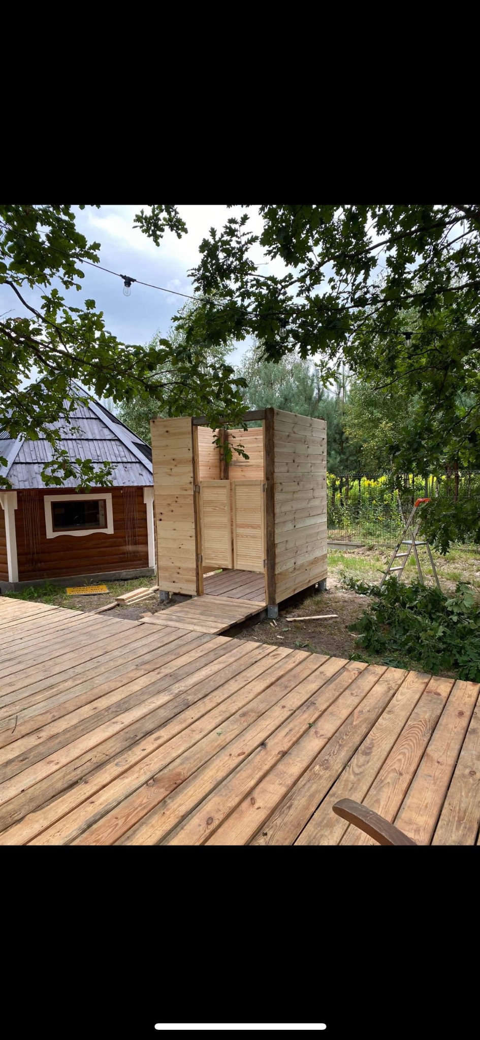 Drewniany taras prowadzący do szatni ogrodowej z surowego drewna, widok na mały domek i zieleń w tle.