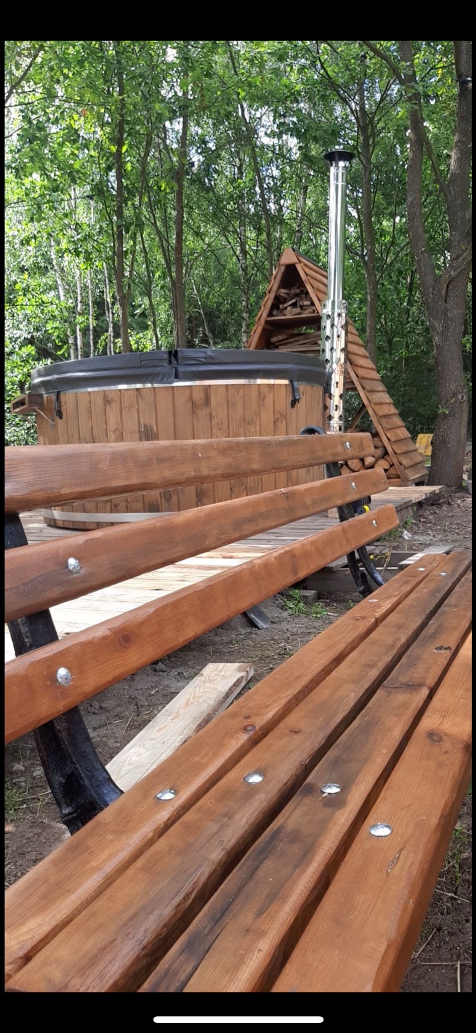 Drewniana ławka z metalowymi nogami na pierwszym planie, w tle okrągła balia z drewnianą obudową i kominem oraz drewutnia w kształcie trójkąta, otoczone zielenią.