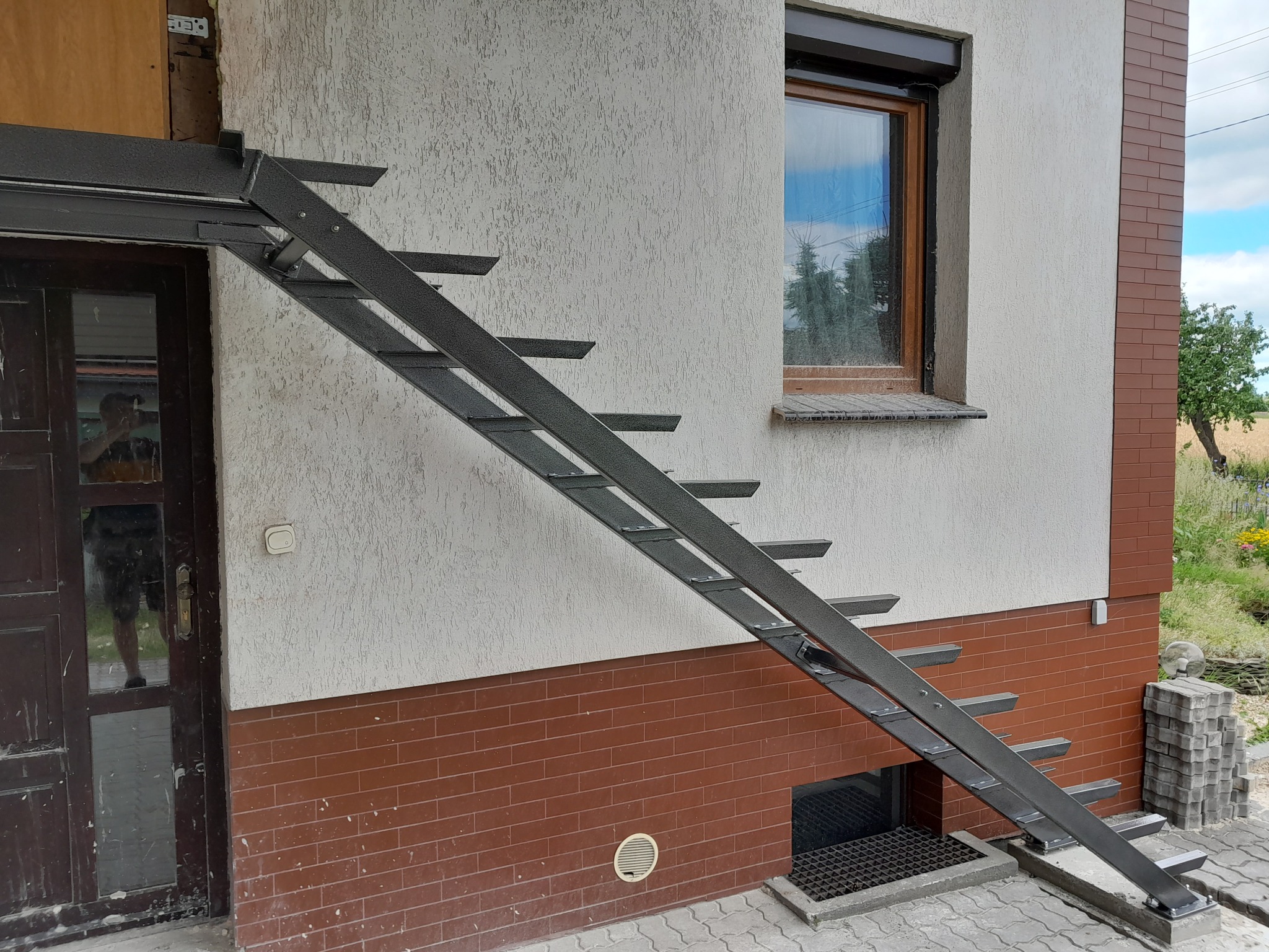 Zewnętrzne metalowe schody przymocowane do ściany domu z brązową cegłą, widoczne okno z brązową ramą i roletą.