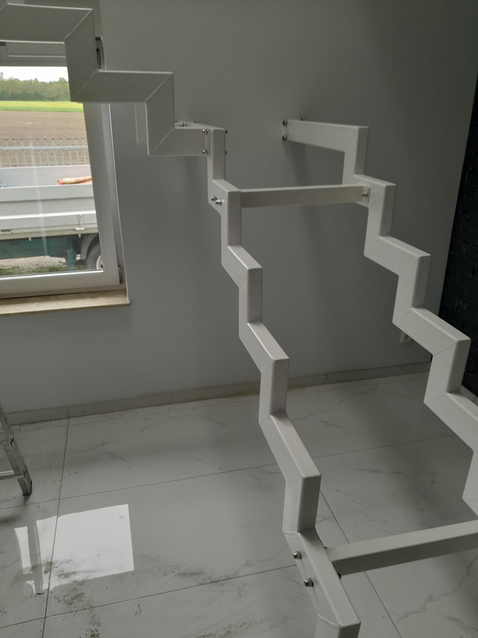 Surowa, metalowa konstrukcja schodów wewnętrznych w trakcie montażu, z widocznym oknem i fragmentem drabiny.