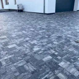 Ułożona kostka brukowa w odcieniach szarości przed nowoczesnym domem z garażem i klimatyzatorem na ścianie.