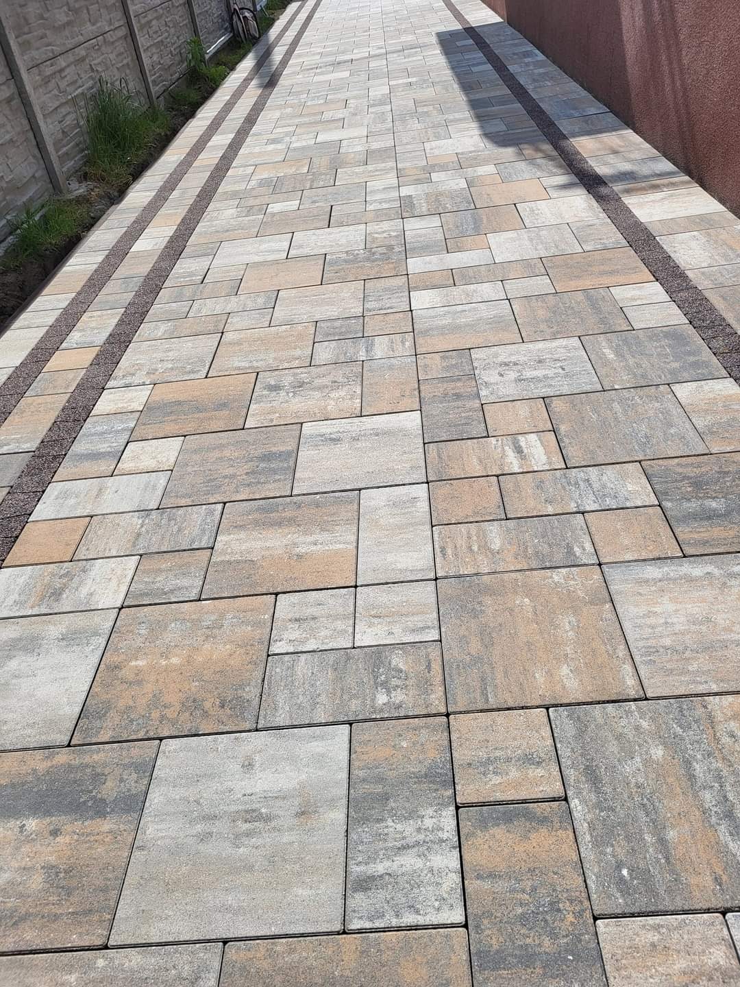 Długa ścieżka z ułożonej kostki brukowej w odcieniach szarości i beżu, oddzielona od trawnika i ściany pasami drobnego brązowego żwiru.
