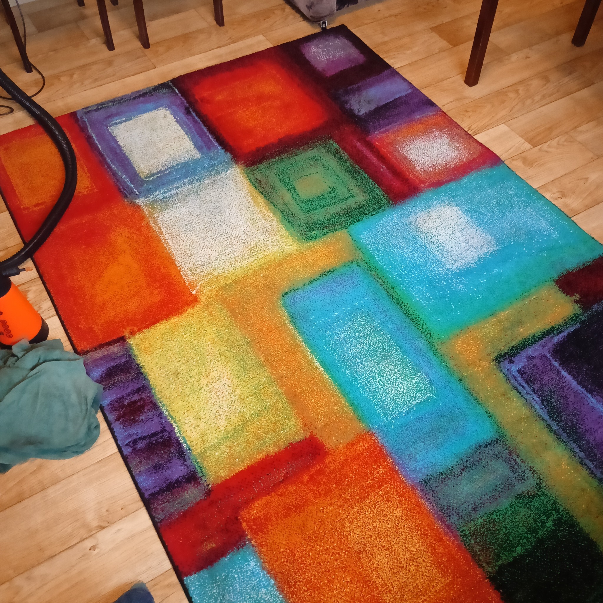 Wielobarwny dywan z geometrycznym wzorem, częściowo mokry po czyszczeniu, z widocznym sprzętem do prania i fragmentem podłogi z paneli.