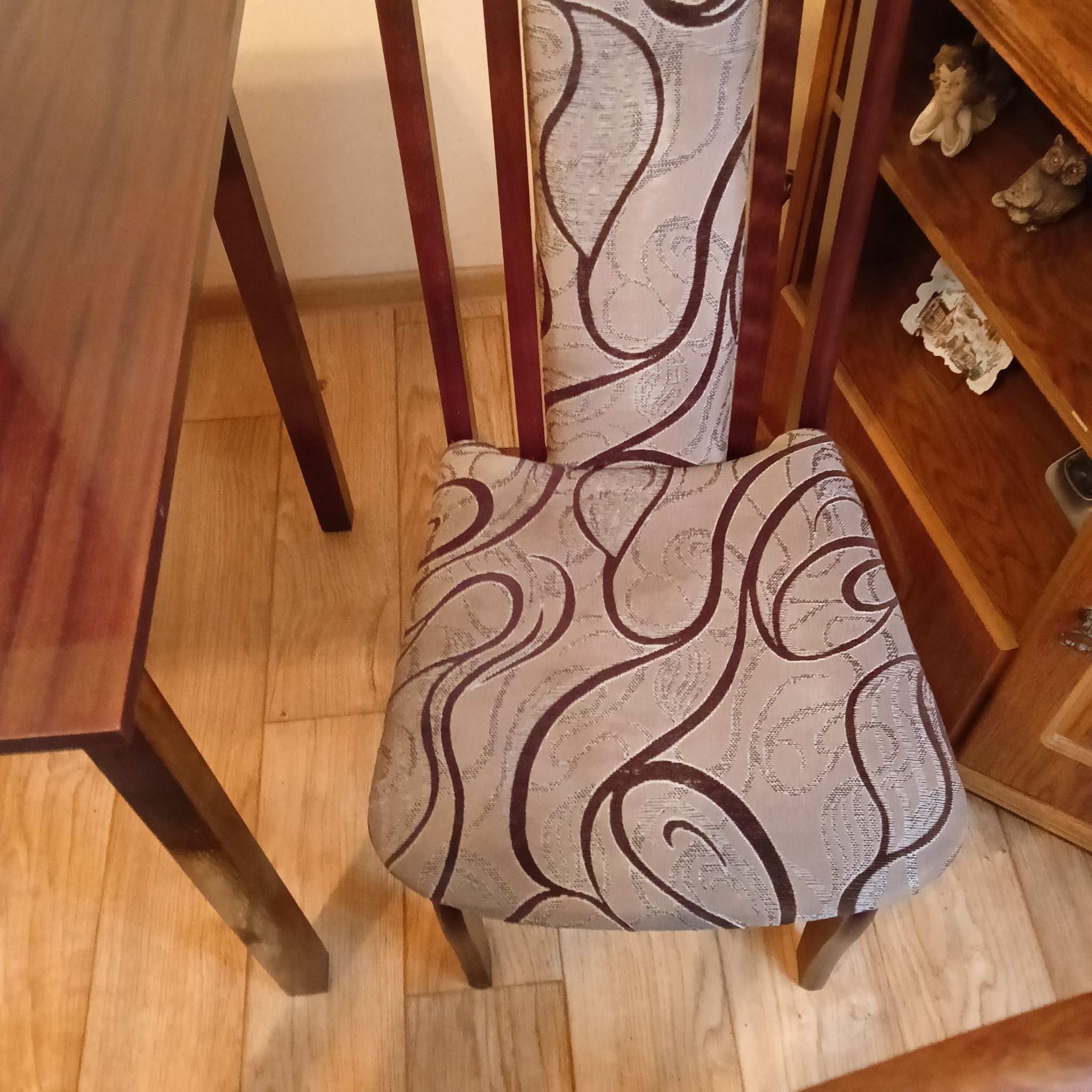 Krzesło z tapicerowanym siedziskiem i oparciem w kremowo-brązowe wzory, stojące obok drewnianego stołu i szafki z ozdobnymi figurkami.