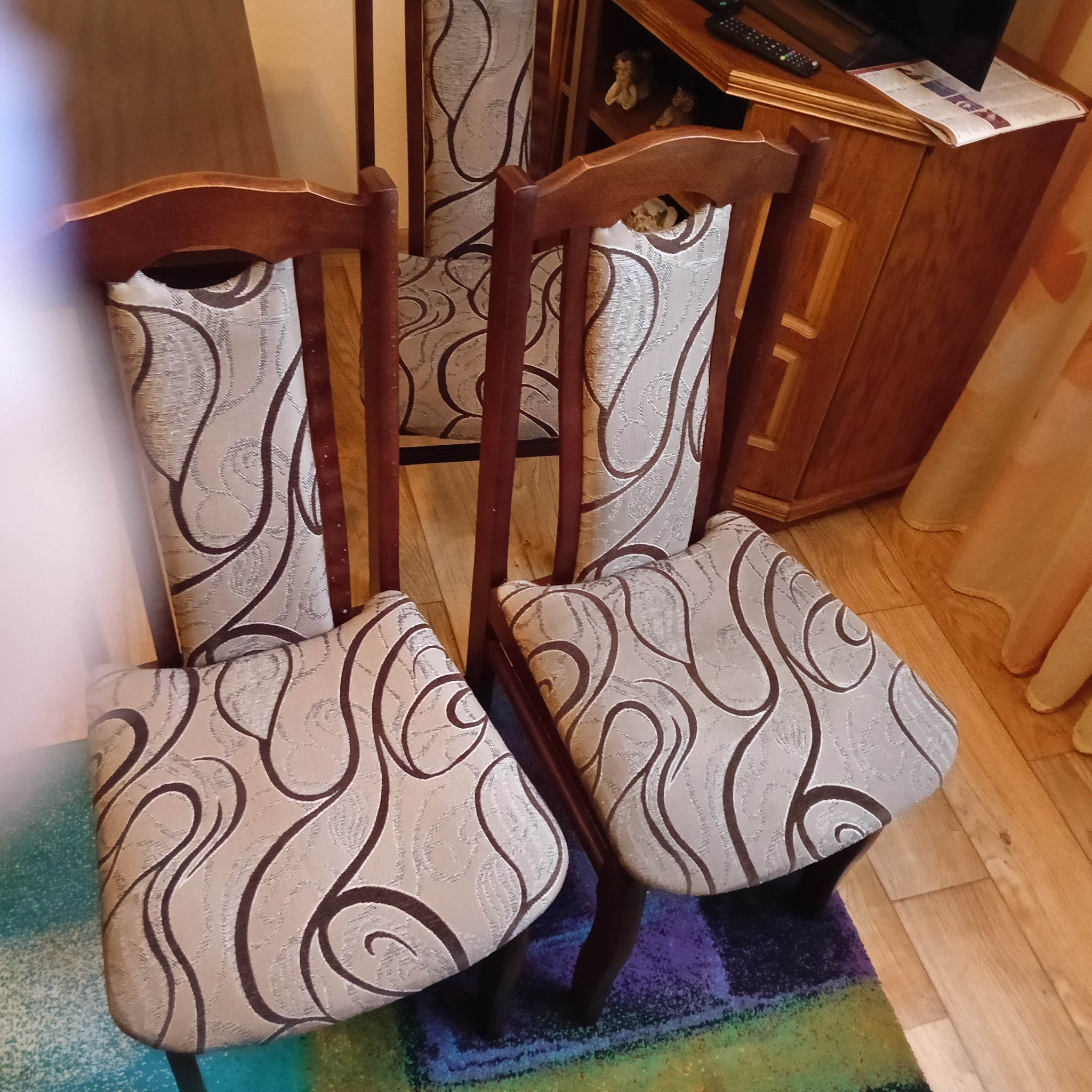 Cztery krzesła z tapicerką w beżowo-brązowe wzory stoją na dywanie z widocznym drewnianym parkietem i komodą w tle.