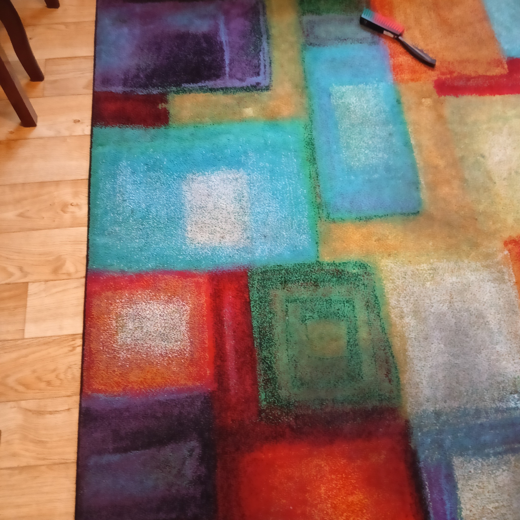 Wielobarwny dywan o geometrycznym wzorze z widoczną szczotką do czyszczenia na drewnianej podłodze.